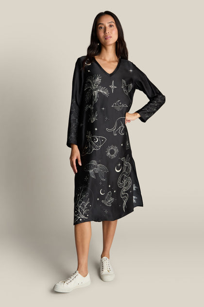 A woman stands wearing the Violet Dress, a black, long-sleeved garment adorned with detailed celestial and nature-themed designs, paired with white sneakers. The deep V-neck adds an elegant touch as she confidently rests one hand on her hip.