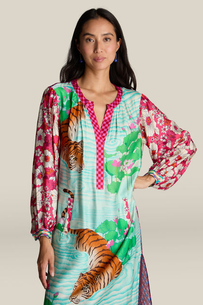 A woman wearing the vibrant Twisted Love Hanna Placket Dress, adorned with a breathtaking mix of tiger illustrations and floral designs, stands against a plain background.