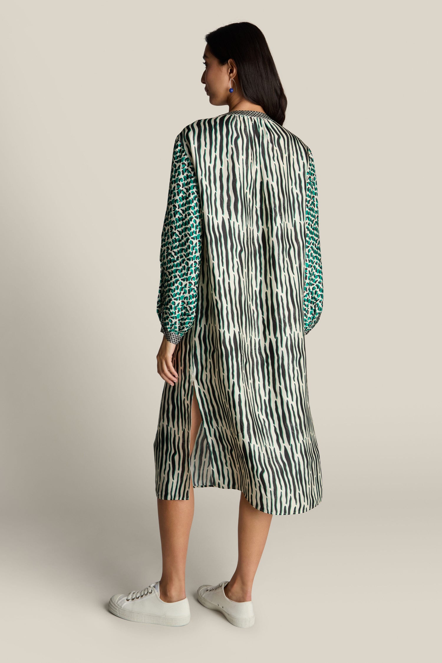 A person stands in a Tropical Hanna Placket Dress (ME369), featuring vibrant green, black, and white stripes interspersed with dots. They are viewed from the back while wearing white sneakers and facing slightly to the side, exuding a contemporary edge.