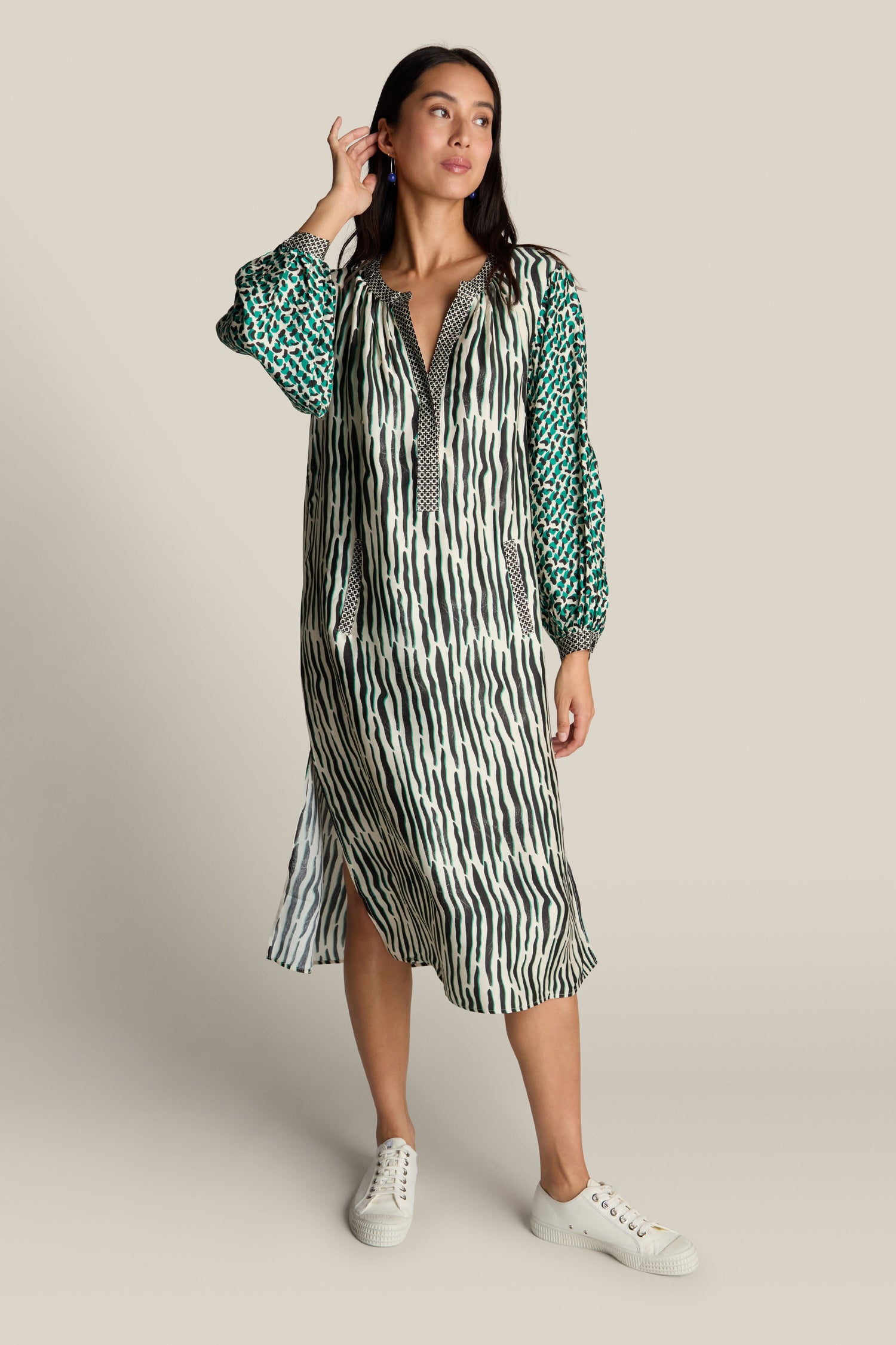 Against a beige background, a woman showcases the Tropical Hanna Placket Dress from ME369. This long, patterned statement dress boasts green and black stripes complemented by abstract print sleeves, paired with white sneakers. She has one hand raised to her ear.