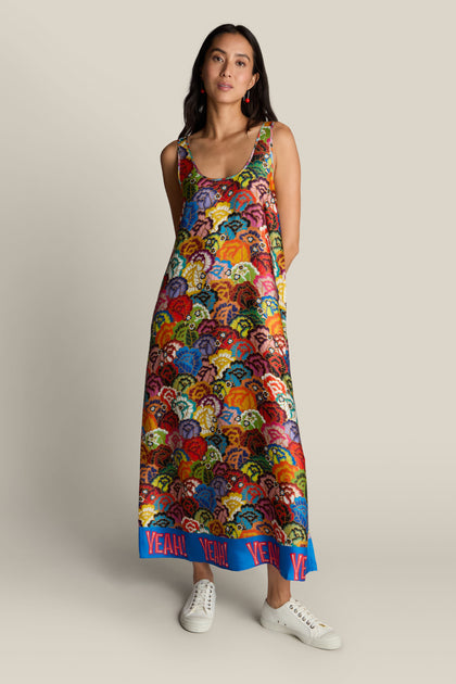 A woman stands against a neutral background, wearing the Allison Dress, which boasts a sleeveless and vibrant floral design adorned with the word "YEAH!" on the blue hem. She pairs it with white sneakers.