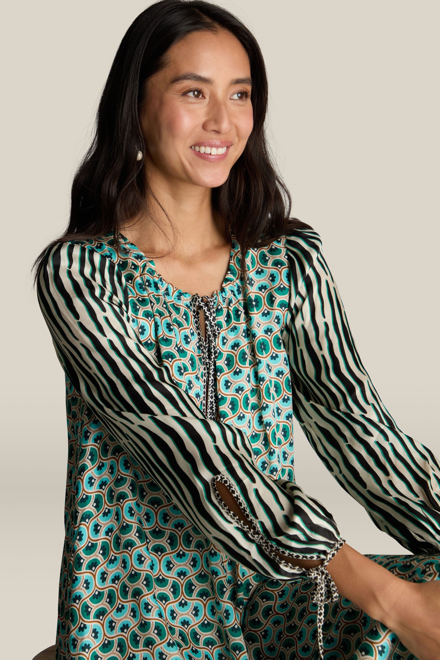 A woman with long dark hair, wearing a green and black patterned blouse, smiles while looking slightly to the side, exuding the vibrant aesthetic of a Polette Dress.