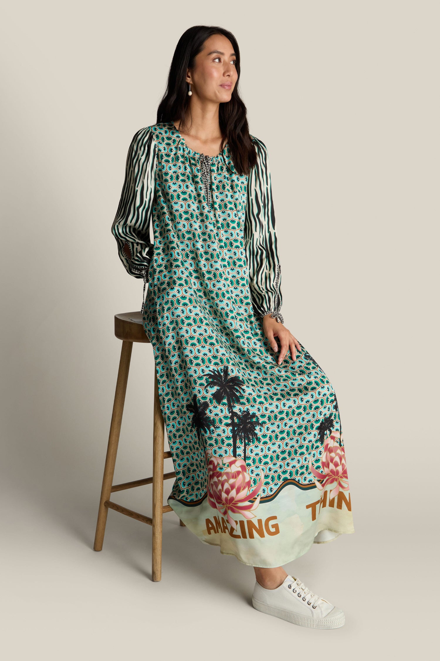 A woman with long dark hair sits on a wooden stool, wearing a long, patterned Polette Dress by ME369 with "Amazing Things" written at the hem. She looks to her right and complements her outfit with white sneakers.