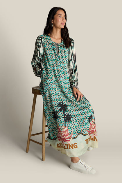 A woman with long dark hair sits on a wooden stool, wearing a long, patterned Polette Dress by ME369 with "Amazing Things" written at the hem. She looks to her right and complements her outfit with white sneakers.