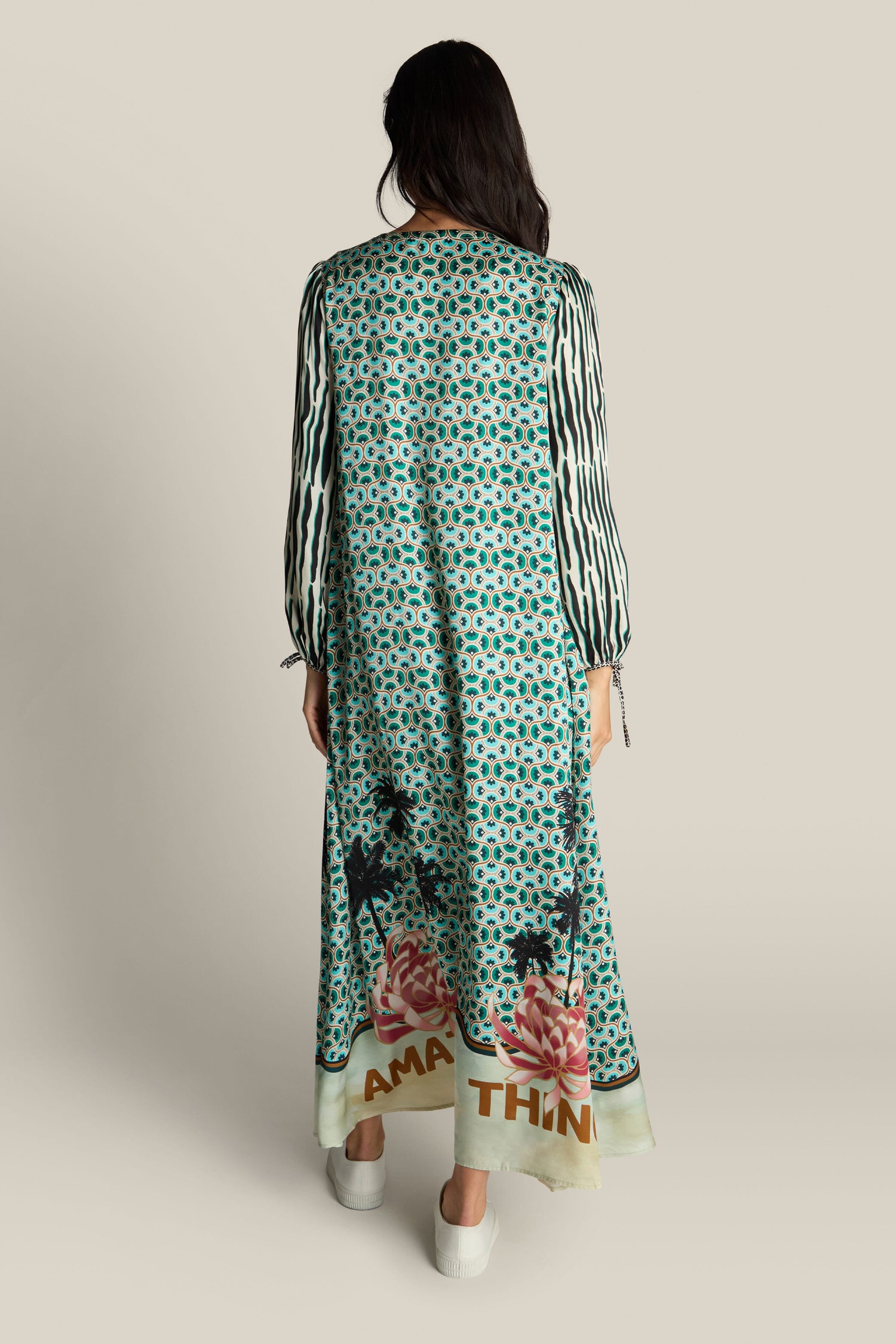 A person with long hair, facing away, is wearing a Polette Dress with a vibrant floral and palm tree pattern near the hem, green fabric, black and white striped sleeves, and white shoes.