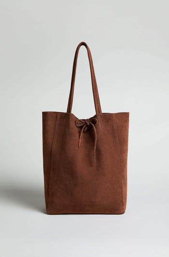 The Soft Suede Tote Bag, a brown minimalist tote with two handles and a front tie detail, is shown upright against a plain light background.