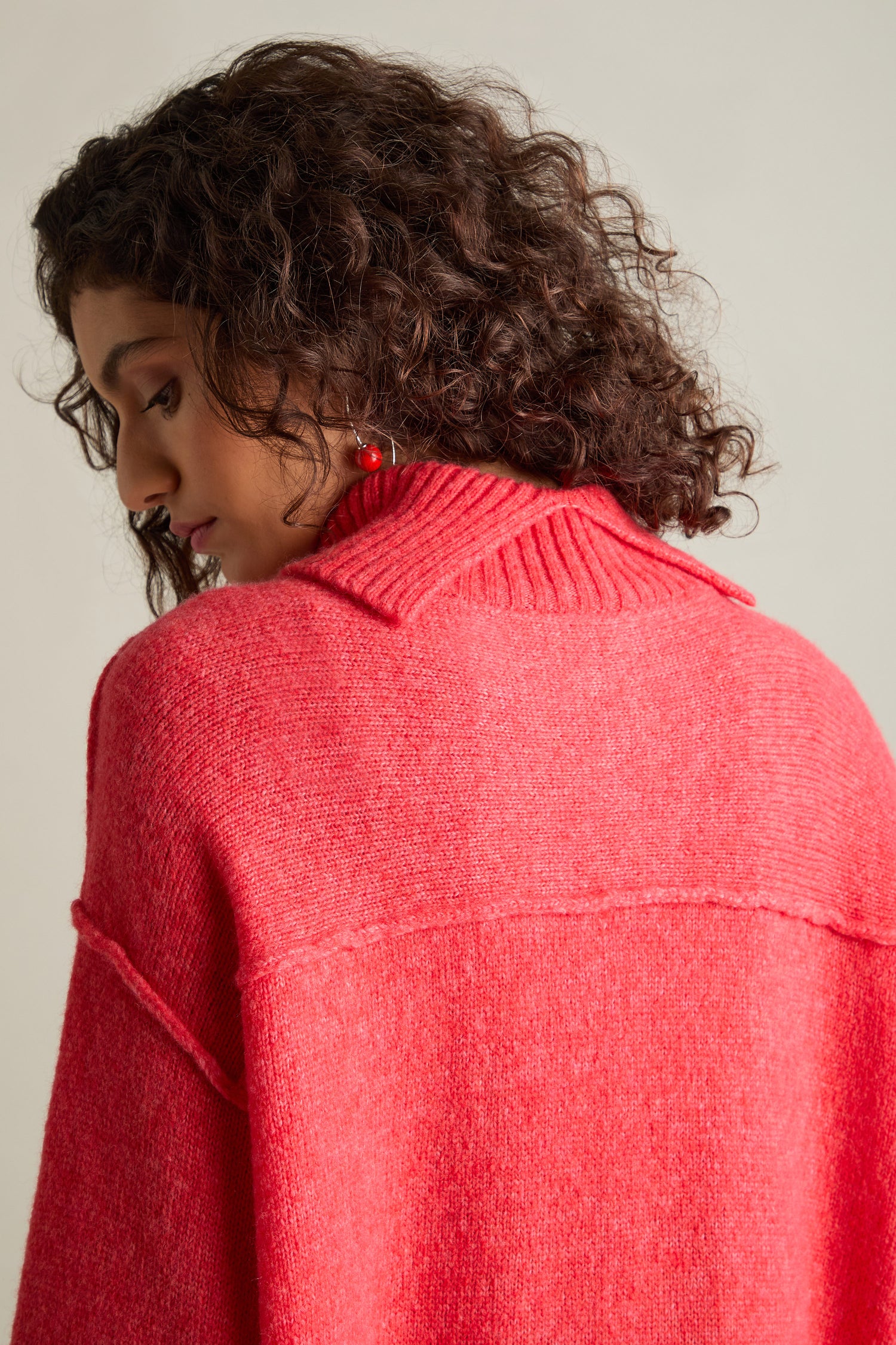 A person with curly hair is seen from the side and back, wearing the Charming Merino Polo Jumper in a striking red color, showcasing its delightful cable knit detailing. They appear to be looking downwards.