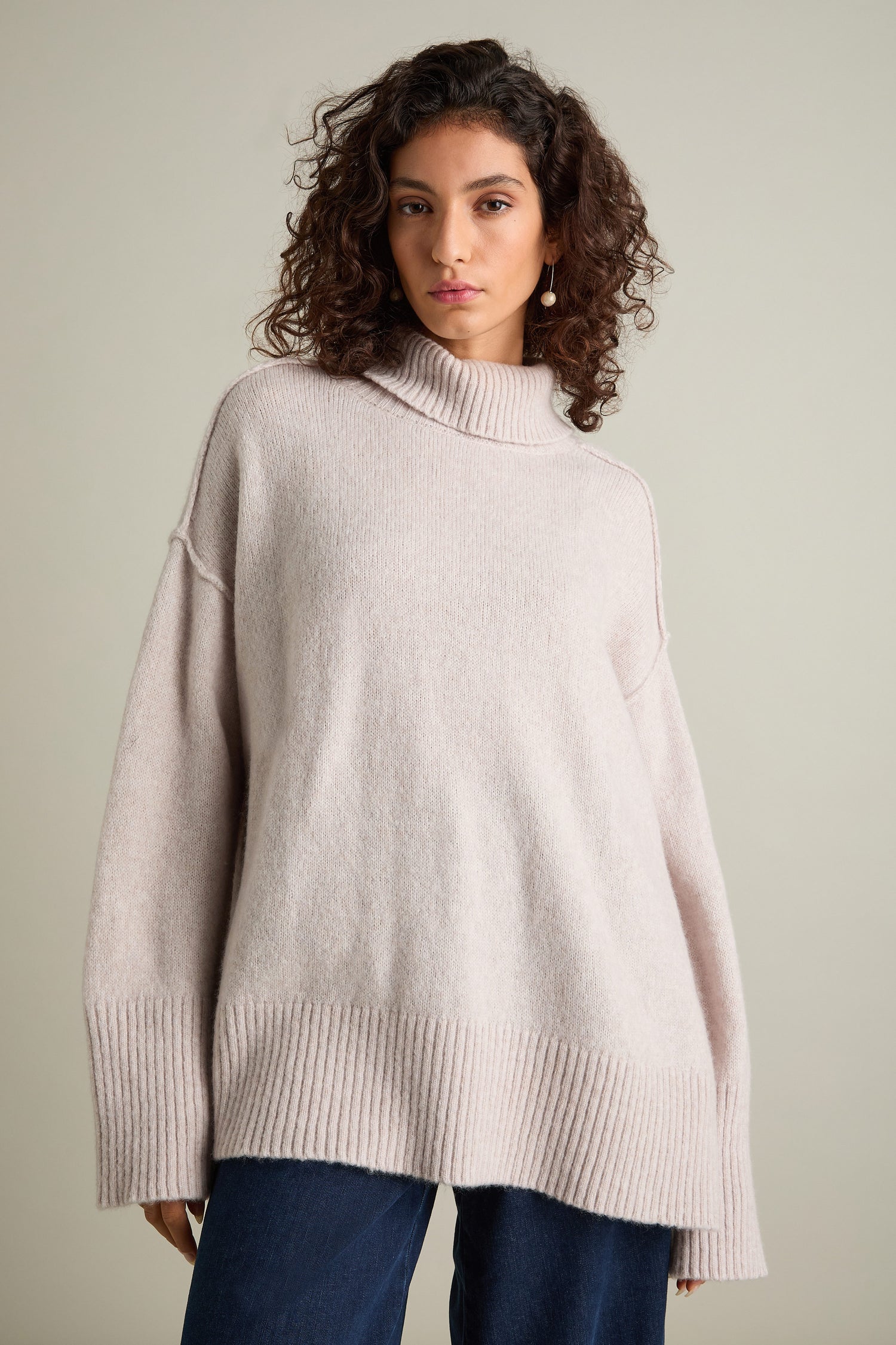 A person wearing an oversized "Charming Merino Polo Jumper" in beige, crafted from extrafine merino wool, and blue jeans stands against a plain neutral background.