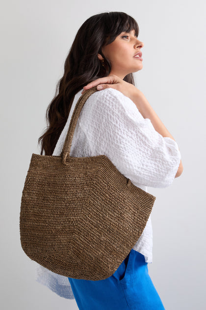 A woman in a white textured blouse and blue pants holds the Handwoven Double Crochet Raffia Basket tote over her shoulder, standing against a plain, light-colored background.