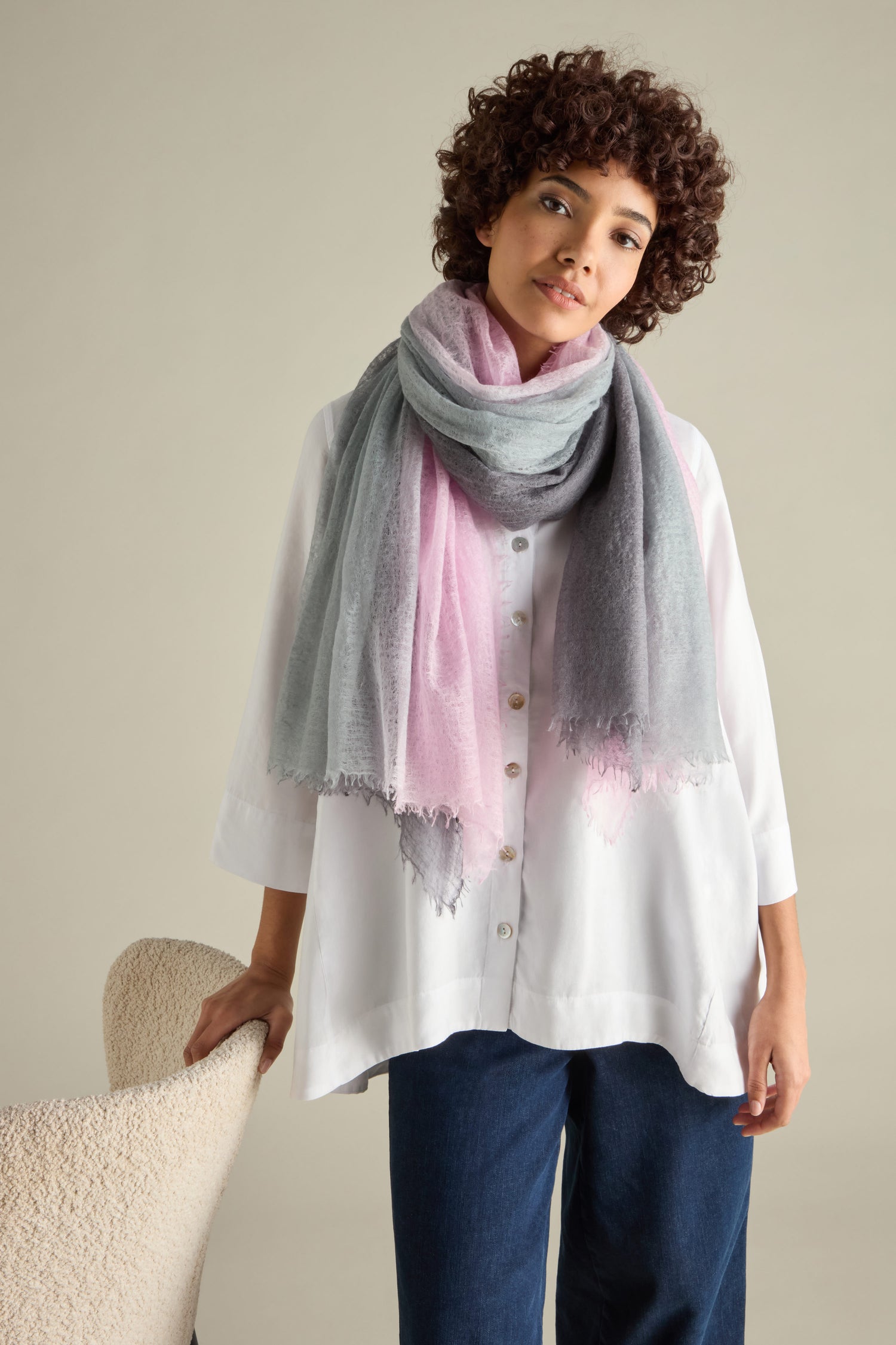 A person wearing a white button-up shirt and blue pants stands next to a beige chair, draped in the St Tropez Cashmere Shawl—a pink and gray handcrafted piece from Nepal.