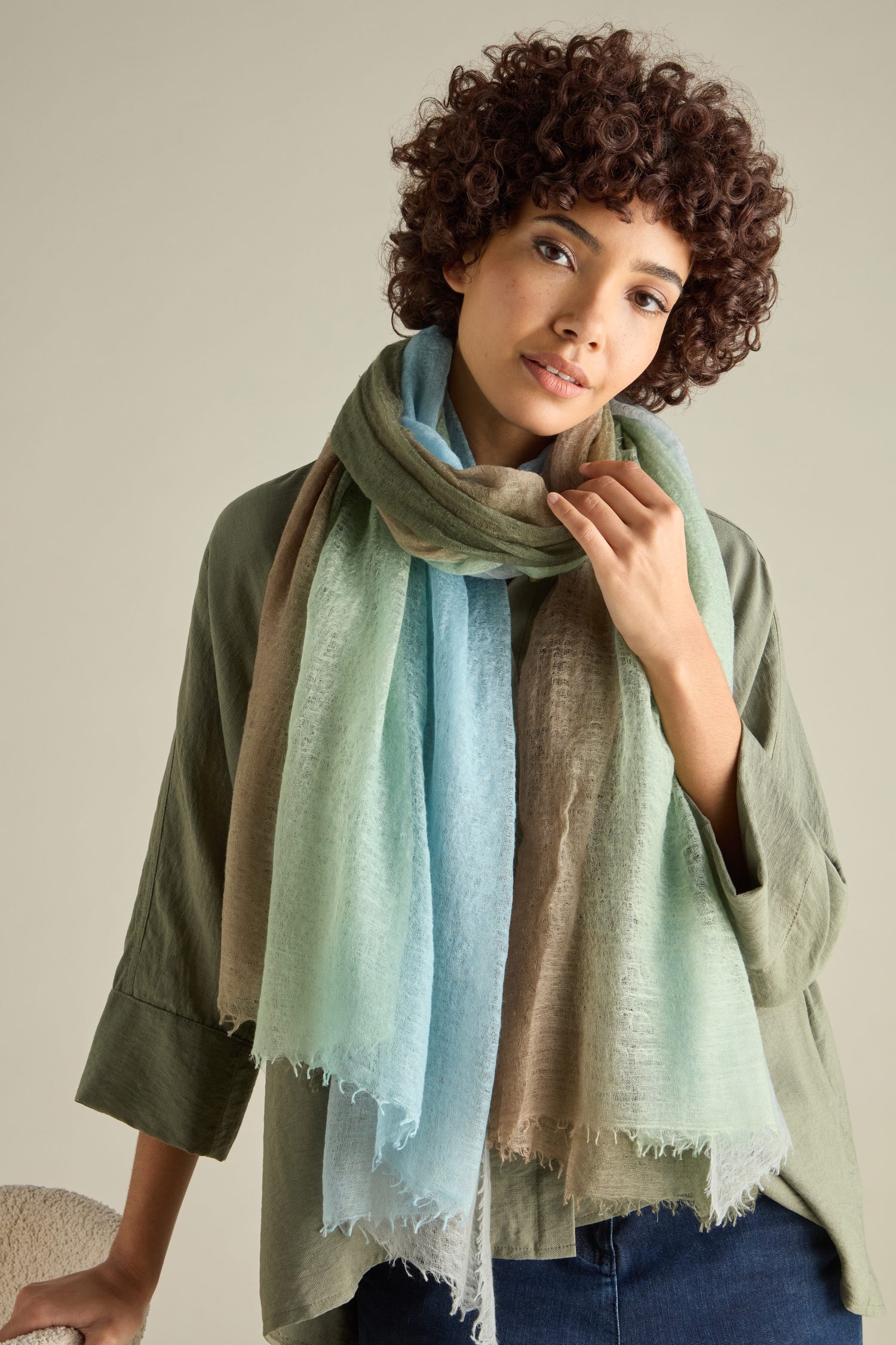 A person with curly hair wears a green shirt and a large, St Tropez Cashmere Shawl handcrafted in Nepal, standing against a plain background.