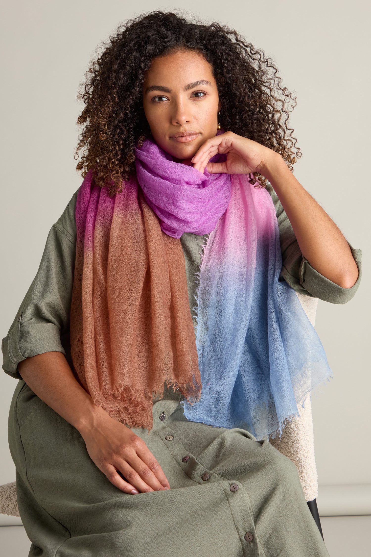 A person with curly hair wears a green buttoned dress and a luxurious St Tropez Cashmere Shawl in shades of pink, brown, and blue, seated against a neutral background.