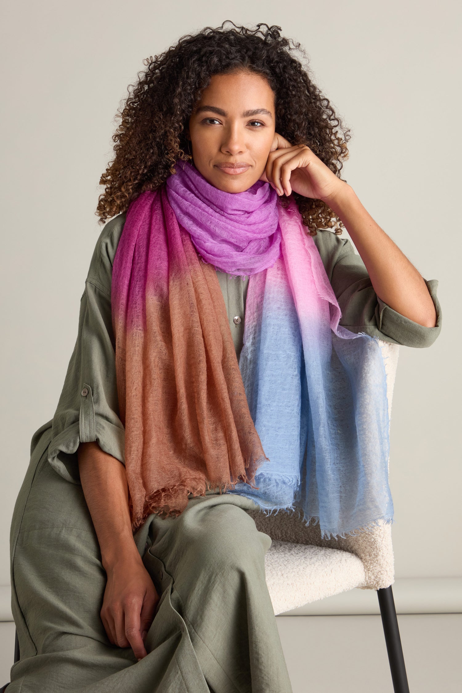 A person with curly hair wearing a green outfit and a St Tropez Cashmere Shawl, featuring a gradient watercolour design, is sitting on a white chair against a plain background.