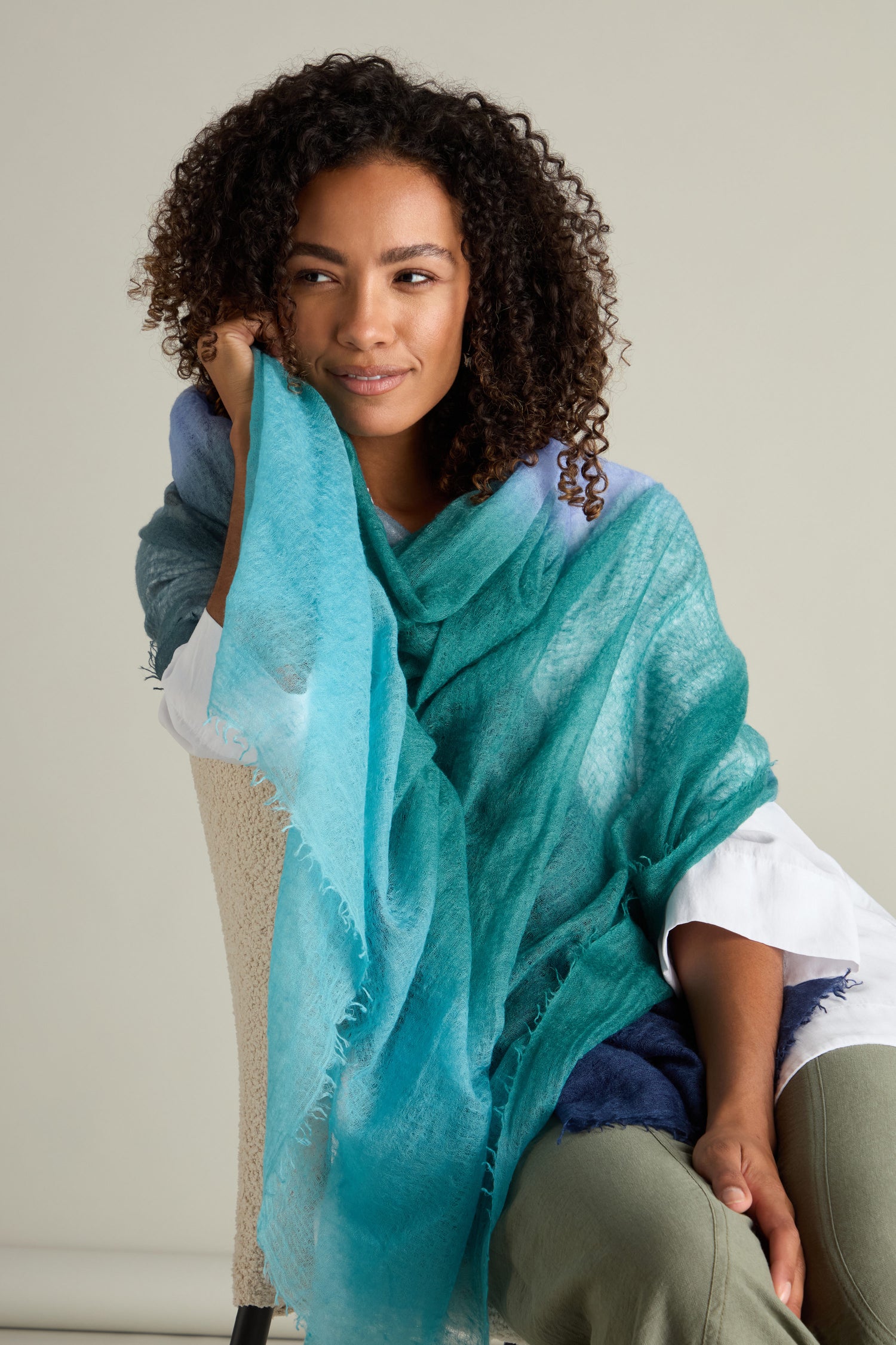 A person with curly hair sits on a chair, draped in a luxurious St Tropez Cashmere Shawl with a gradient watercolour design, looking to the side with a slight smile.