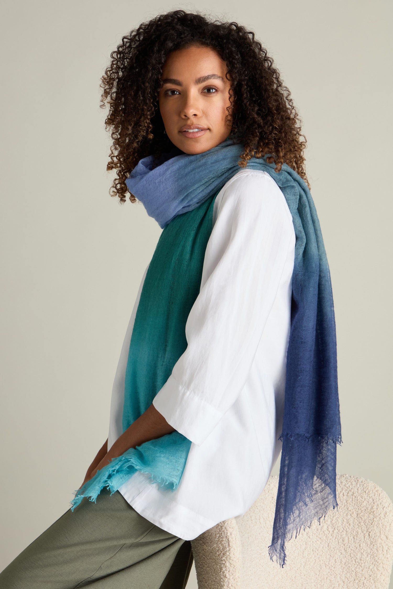 A person with curly hair is sitting, wearing a white long-sleeve shirt, green pants, and a luxurious accessory—a St Tropez Cashmere Shawl featuring a blue-green ombre gradient watercolour design. They look forward with a neutral expression.