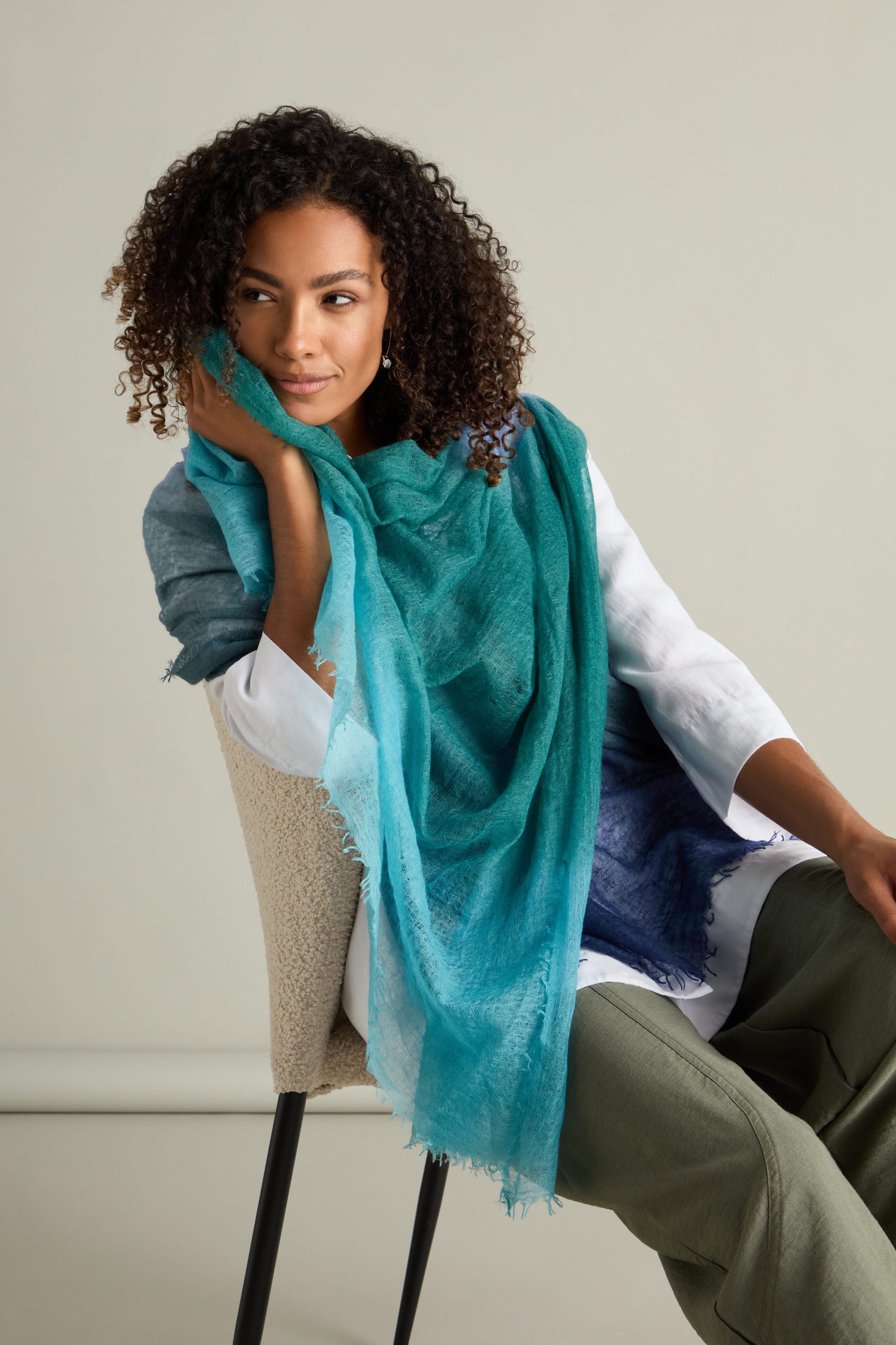 A person with curly hair is sitting on a chair, wearing a white top and green pants, accessorized with the luxurious St Tropez Cashmere Shawl. The shawl features a gradient watercolor design in blue and turquoise, adding an air of elegance as they lean slightly and rest their head on their hand.