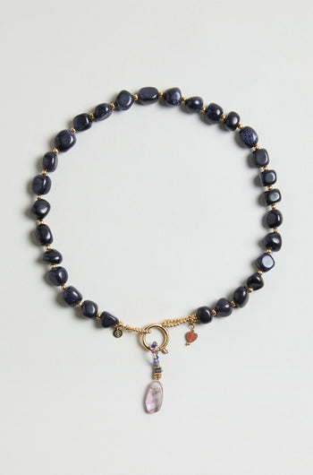 The Sunstone Bead Necklace (MJU33026MG47) features dark blue oval beads, gold accents, a circular gold clasp, small gold and heart charms, and a purple gemstone pendant.