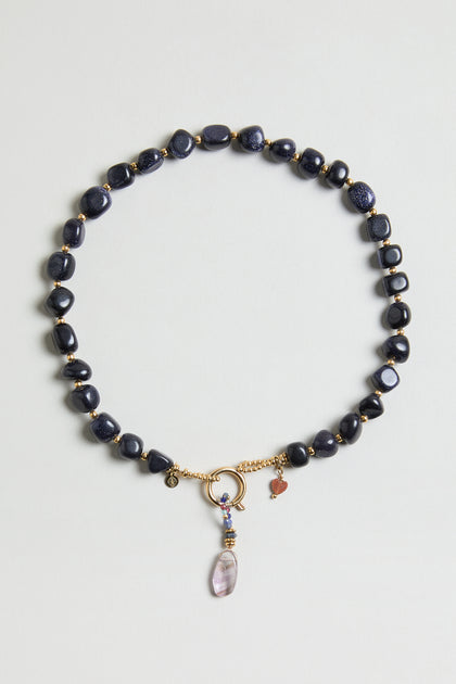 The Sunstone Bead Necklace (MJU33026MG47) features dark blue oval beads, gold accents, a circular gold clasp, small gold and heart charms, and a purple gemstone pendant.