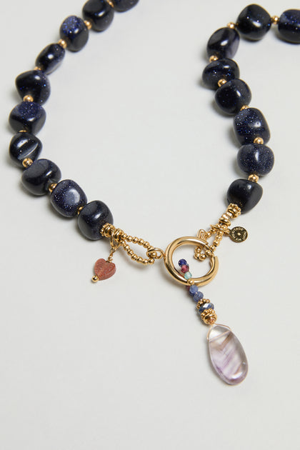 The Sunstone Bead Necklace, MJU33026MG47, features dark blue beads, gold accents with a gold clasp, a small red heart charm, and a pendant showcasing a translucent stone.