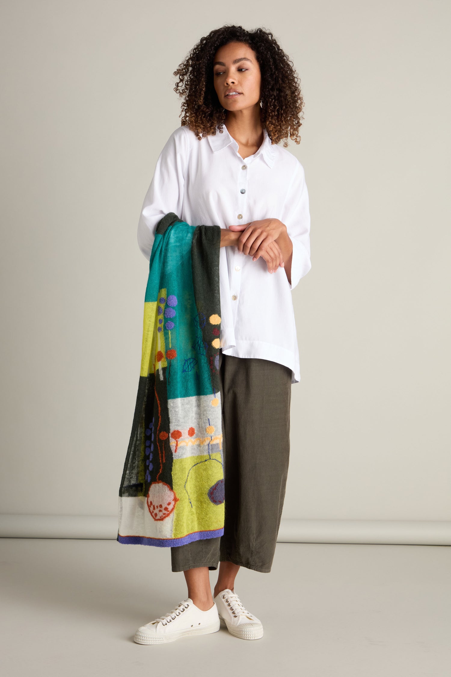 A person with curly hair is standing, holding an Abstract Mohair Shawl— a luxurious shawl adorned with colorful patterns. They are wearing a white button-down shirt, green pants, and white sneakers against a plain beige background.