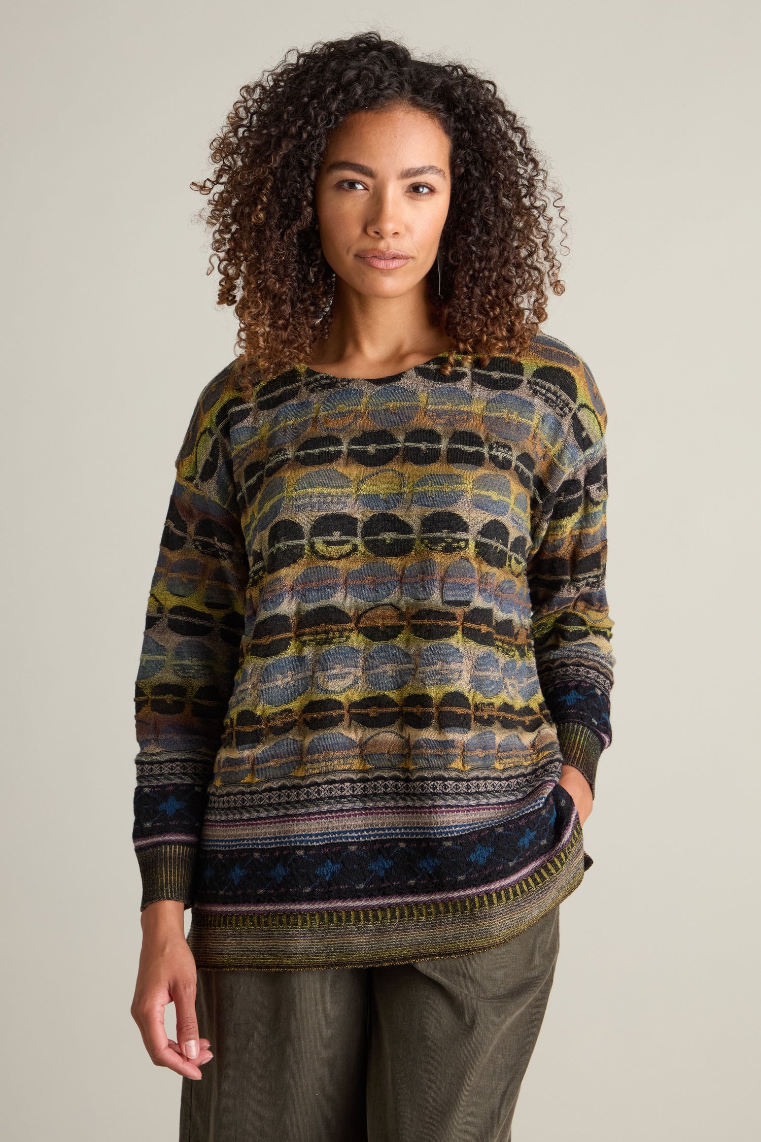 A person with curly hair wears a patterned intarsia knit Round Neck Pullover and green pants, posing in front of a plain background.