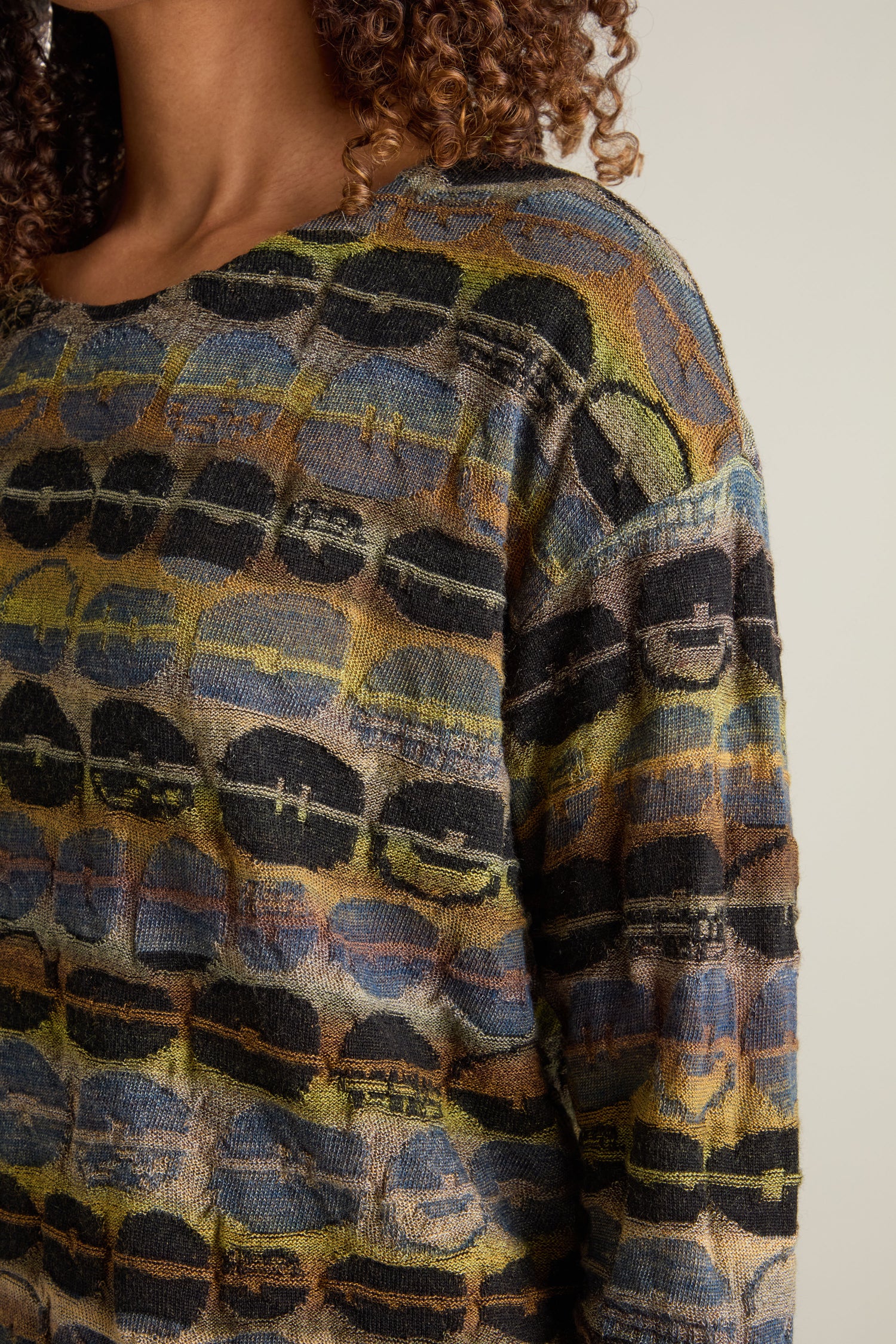 Close-up of a person wearing the Round Neck Pullover, featuring an abstract, multicolored intarsia knit pattern. The image focuses on part of the person's face and upper body, showcasing the artisanal excellence of this sweater.