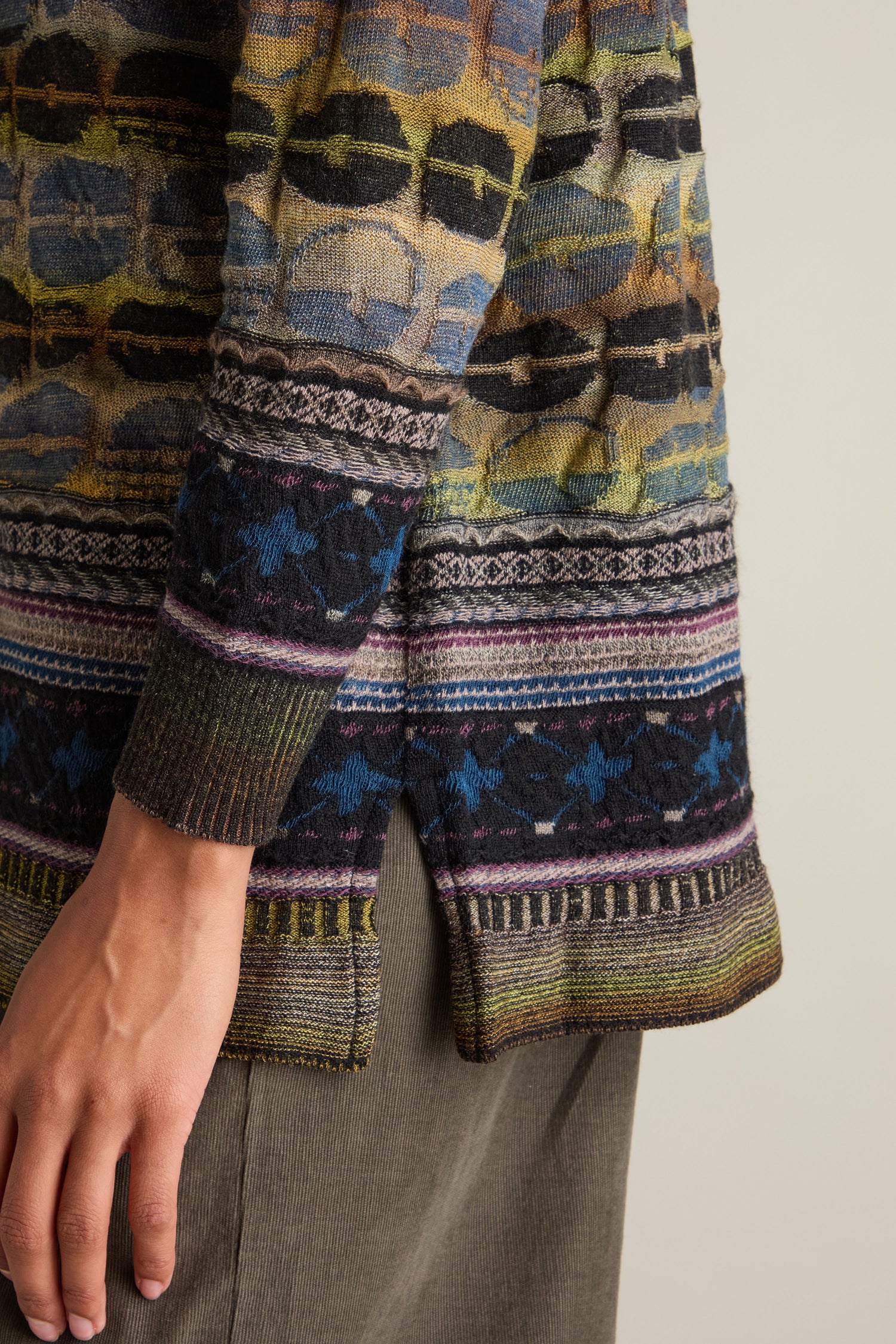 Close-up of a person wearing the Round Neck Pullover, featuring an intarsia knit with a geometric pattern that showcases artisanal excellence. Only the lower part of the pullover and their hand is visible, paired with dark pants.