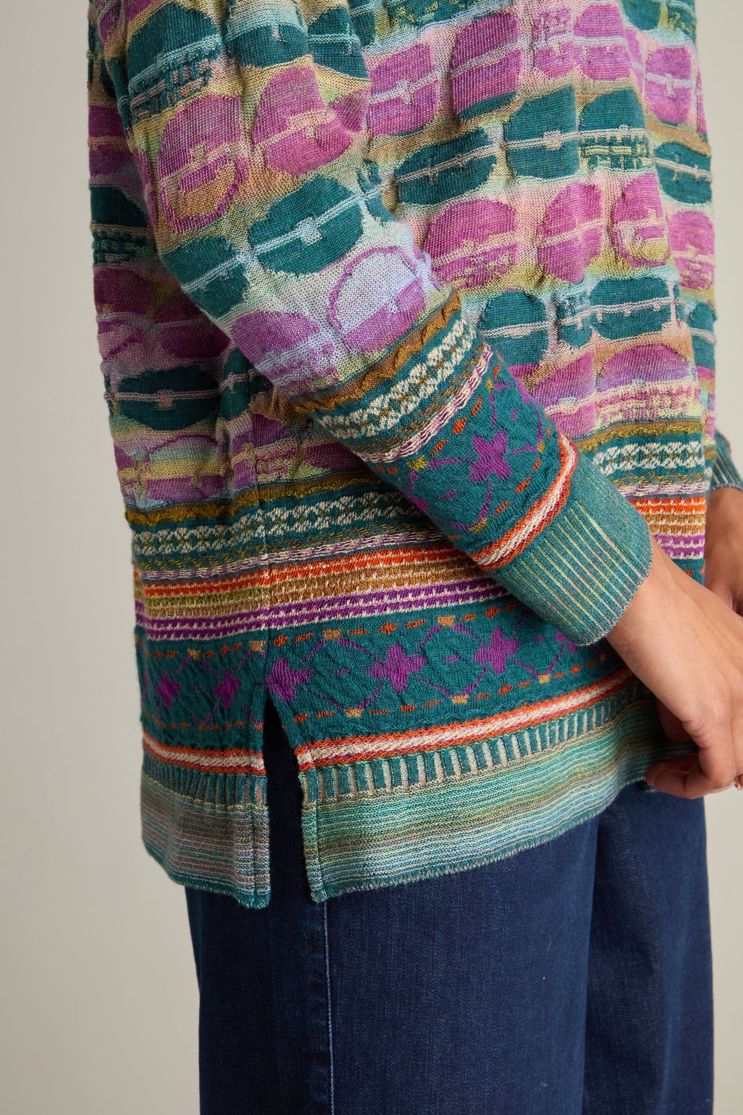 Close-up of a person's torso wearing a vibrant Round Neck Pullover. The intarsia knit sweater showcases intricate patterns in a blend of purples, pinks, greens, and blues. The person's hand rests by their side.