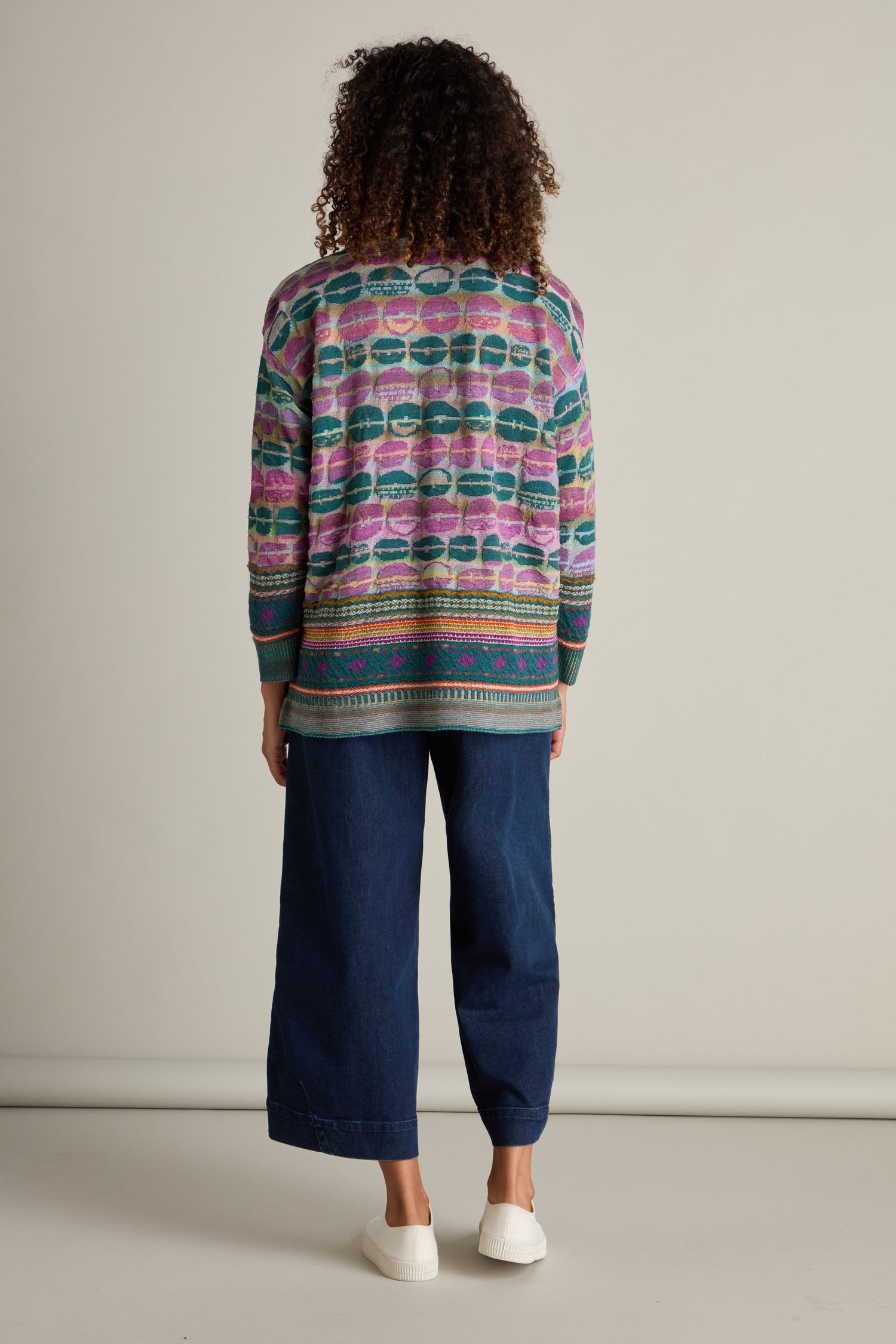 A person with curly hair, seen from the back, is wearing a colorful patterned Round Neck Pullover, dark wide-leg jeans, and white sneakers, standing against a plain background.