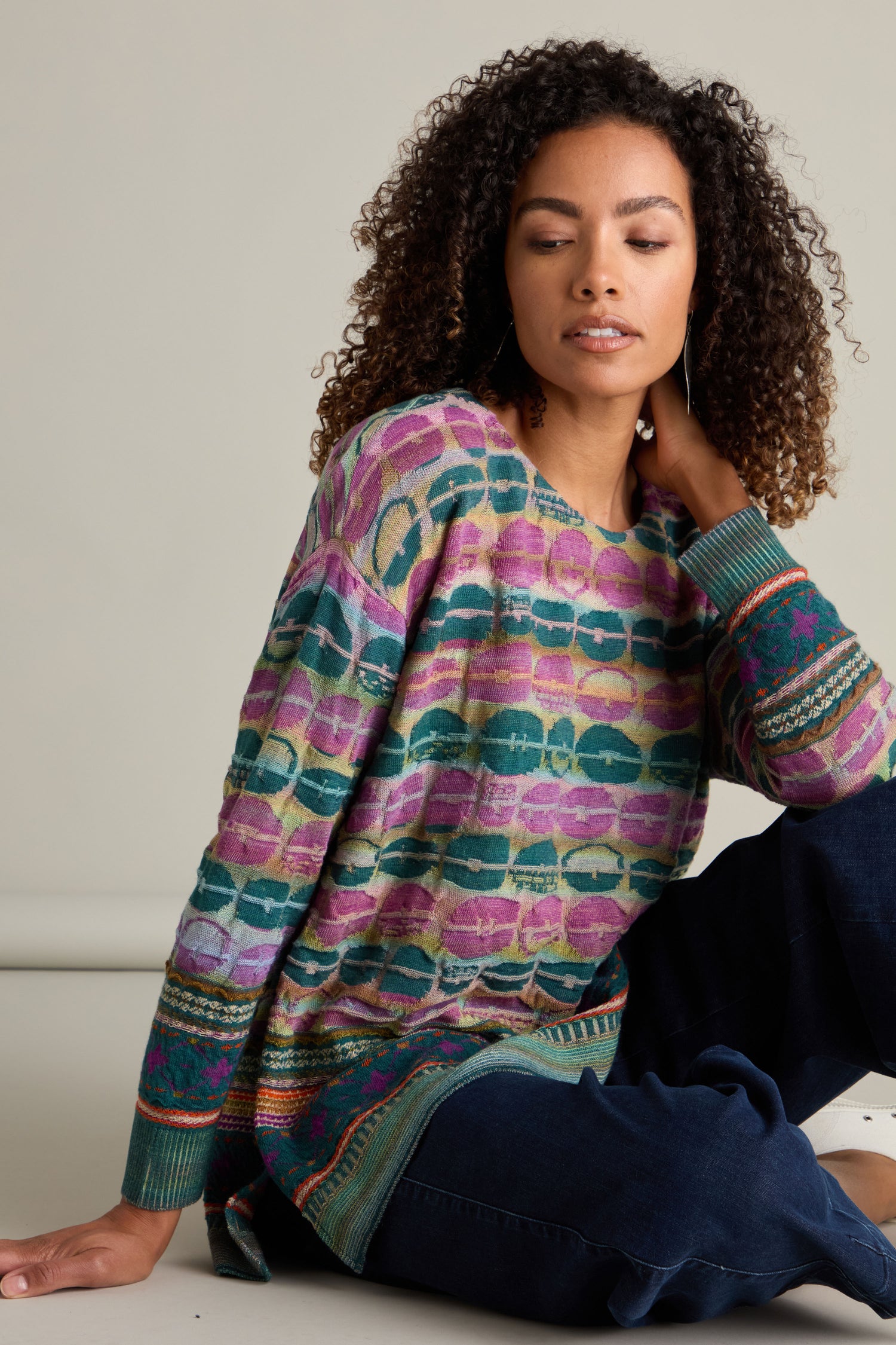 A woman is sitting against a beige background, wearing the colorful Round Neck Pullover, an intarsia knit sweater paired with dark pants, and looking slightly downward. The pullover showcases artisanal excellence in every stitch.