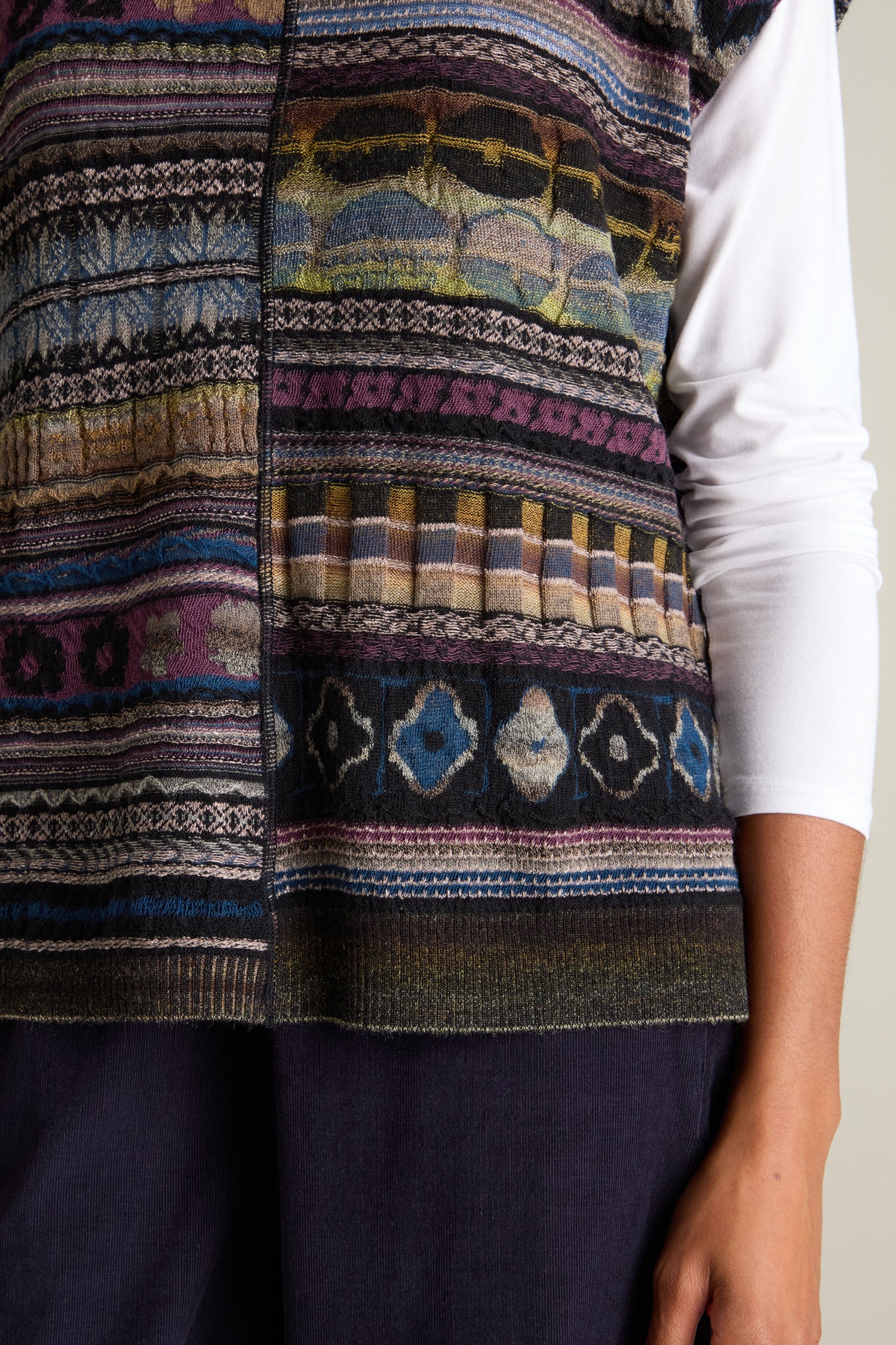 Close-up of a person wearing the Round Neck Tabard by M.&Kyoko, featuring a multi-colored intarsia knit design, over a white long-sleeve shirt, with one arm shown resting by their side.