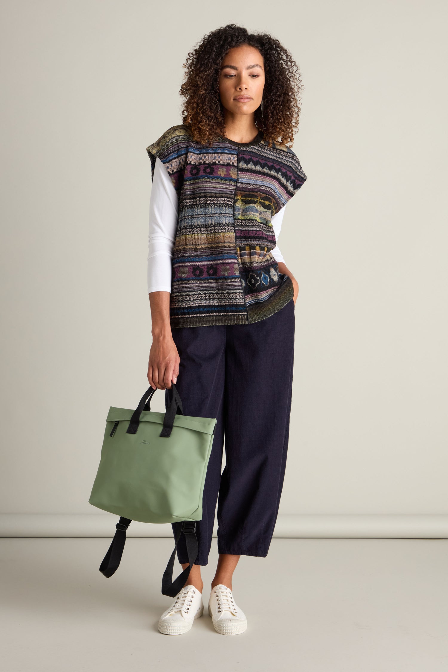 A woman stands against a plain backdrop, wearing a colorful intarsia knit vest over the Round Neck Tabard, dark wide-leg pants, and white sneakers. She's holding a green bag by its straps, showcasing the M.&Kyoko look with effortless style.