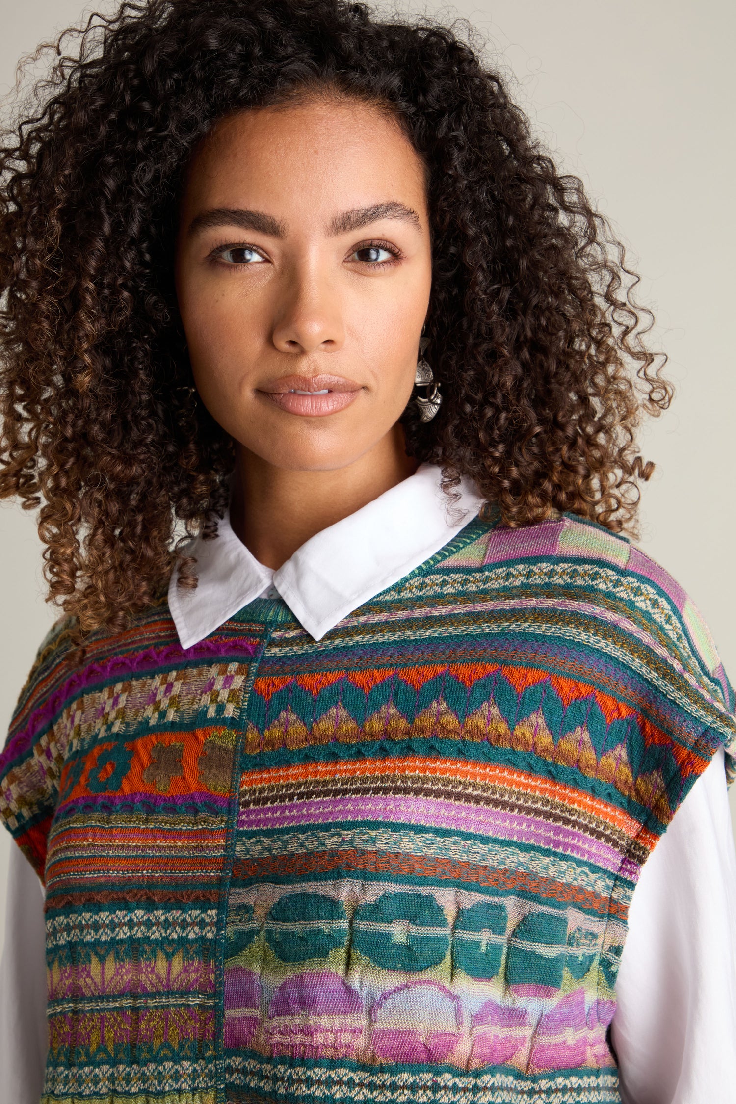 A person with curly hair wears a multicolored, intarsia knit Round Neck Tabard by M.&Kyoko over a white shirt. They are looking directly at the camera.