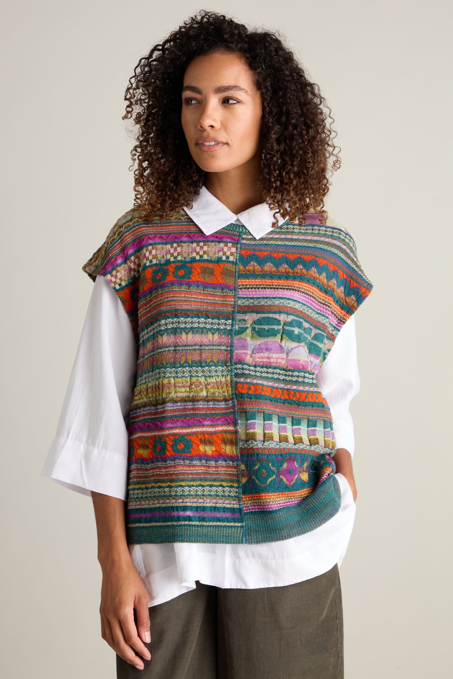 A person is wearing a white button-down shirt under a colorful, Round Neck Tabard by M.&Kyoko. The individual has curly hair and stands against a plain background.