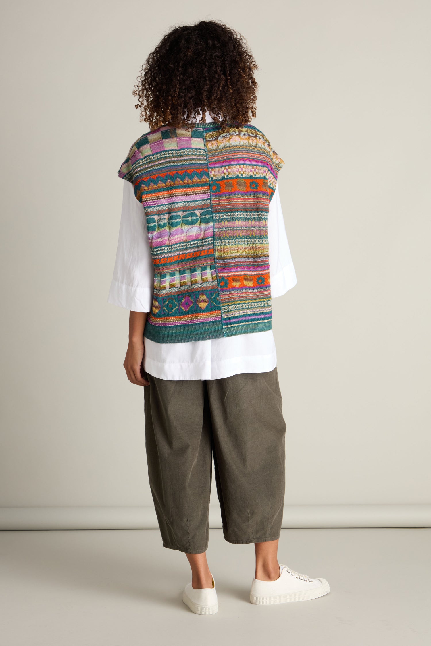 Person in a white long-sleeve shirt, Round Neck Tabard by M.&Kyoko featuring an intarsia knit colorful pattern, olive green pants, and white sneakers, standing with their back to the camera.