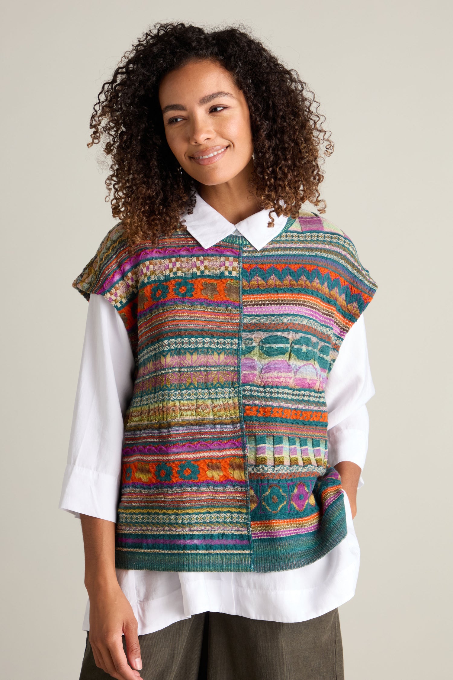 A person with curly hair smiles while wearing a colorful Round Neck Tabard by M.&Kyoko over a white shirt.