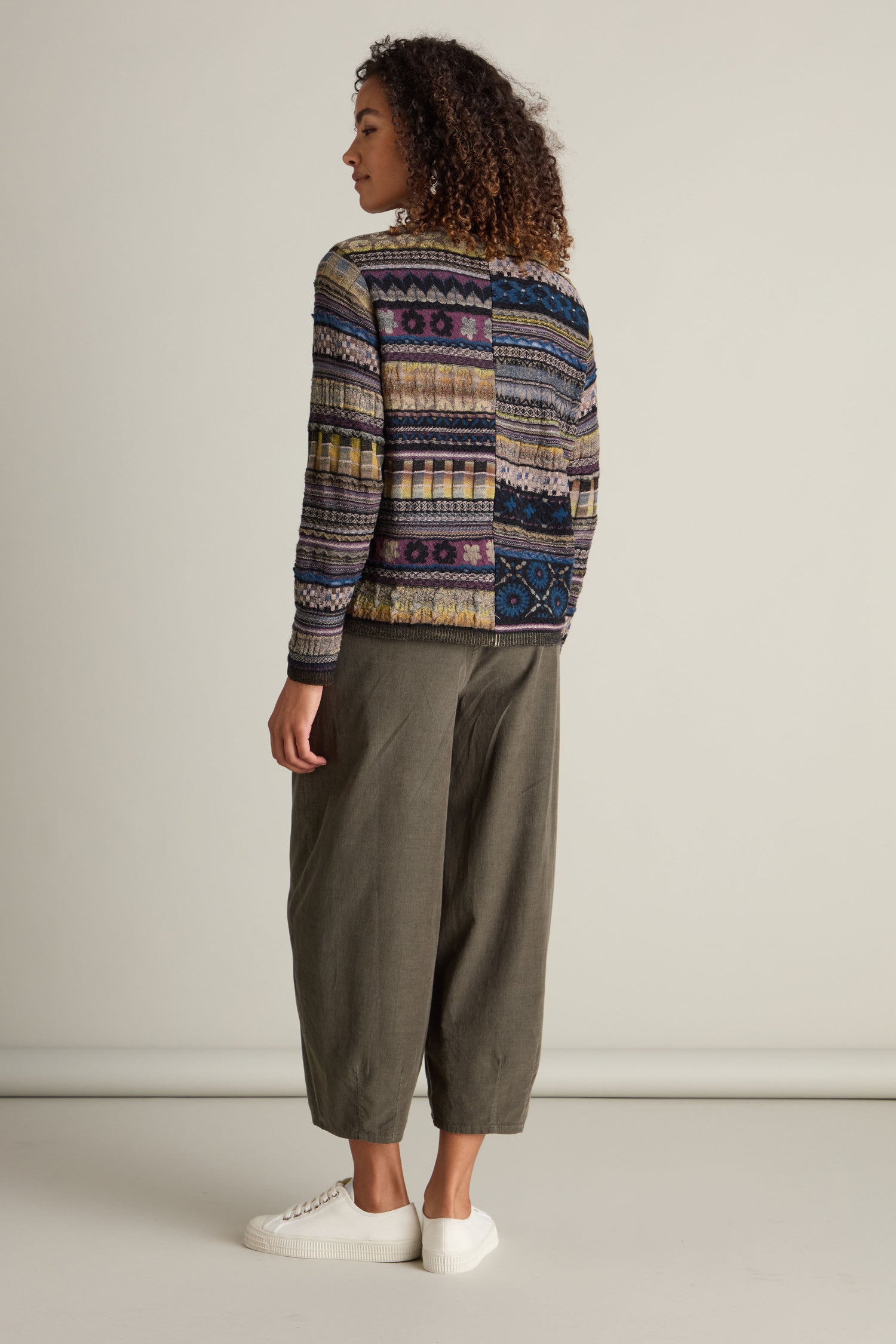 Person with curly hair, wearing a Round Neck Button Cardigan in a colorful intarsia knit design, gray wide-leg pants, and white sneakers, standing and looking to the side against a plain background.