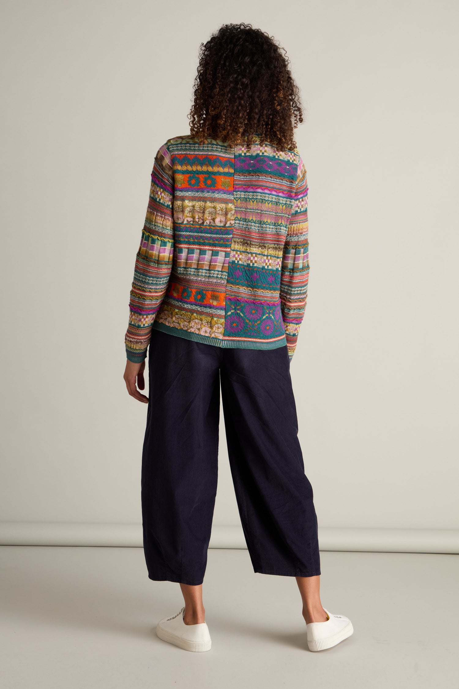 A person with curly hair stands facing away, wearing a colorful Round Neck Button Cardigan featuring an intarsia knit design and dark wide-leg pants, paired with white sneakers. The background is plain and neutral-toned.