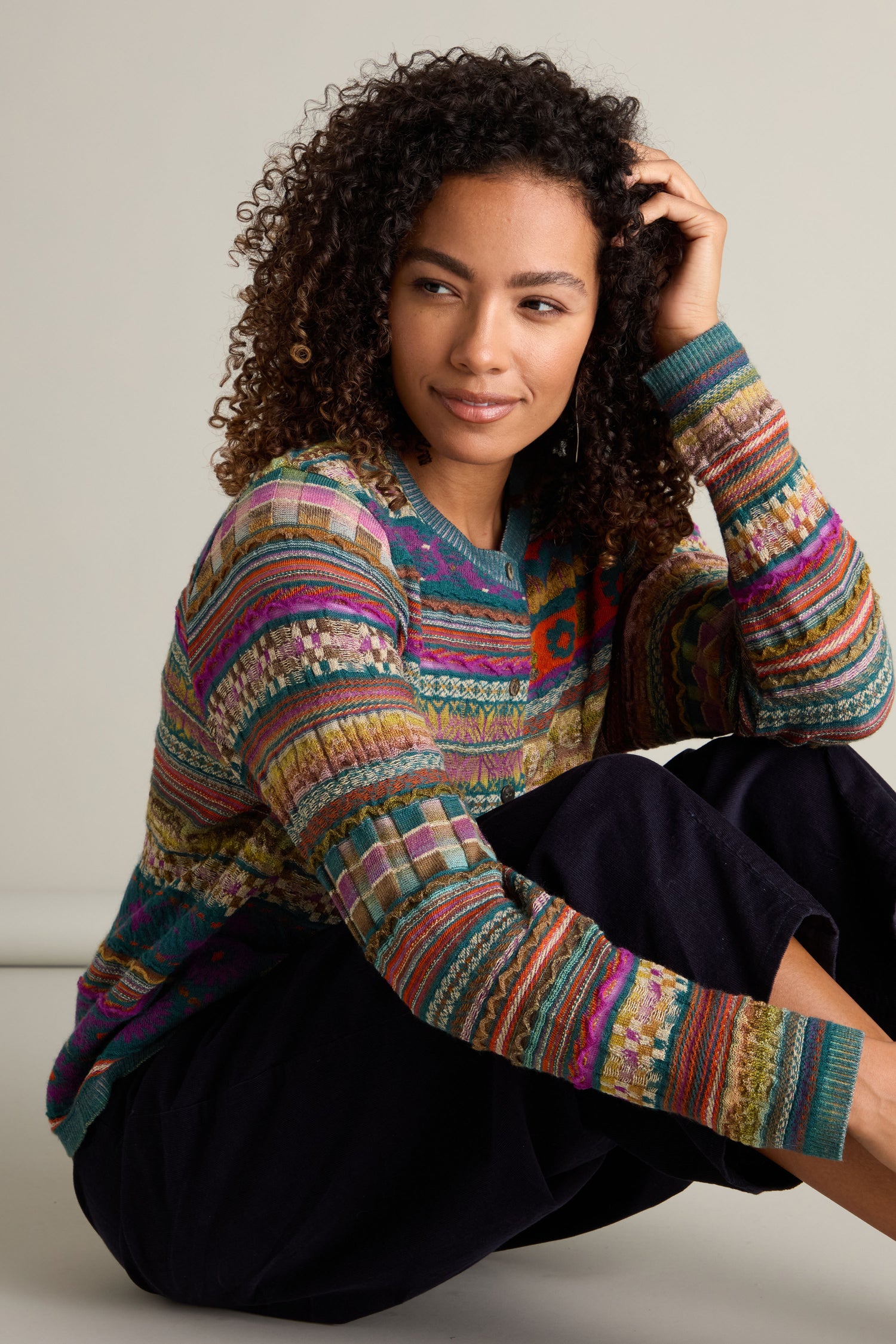 A person with curly hair sits cross-legged, resting one hand on their head. They're wearing dark pants and a colorful patterned Round Neck Button Cardigan showcasing an intarsia knit design, looking off to the side.