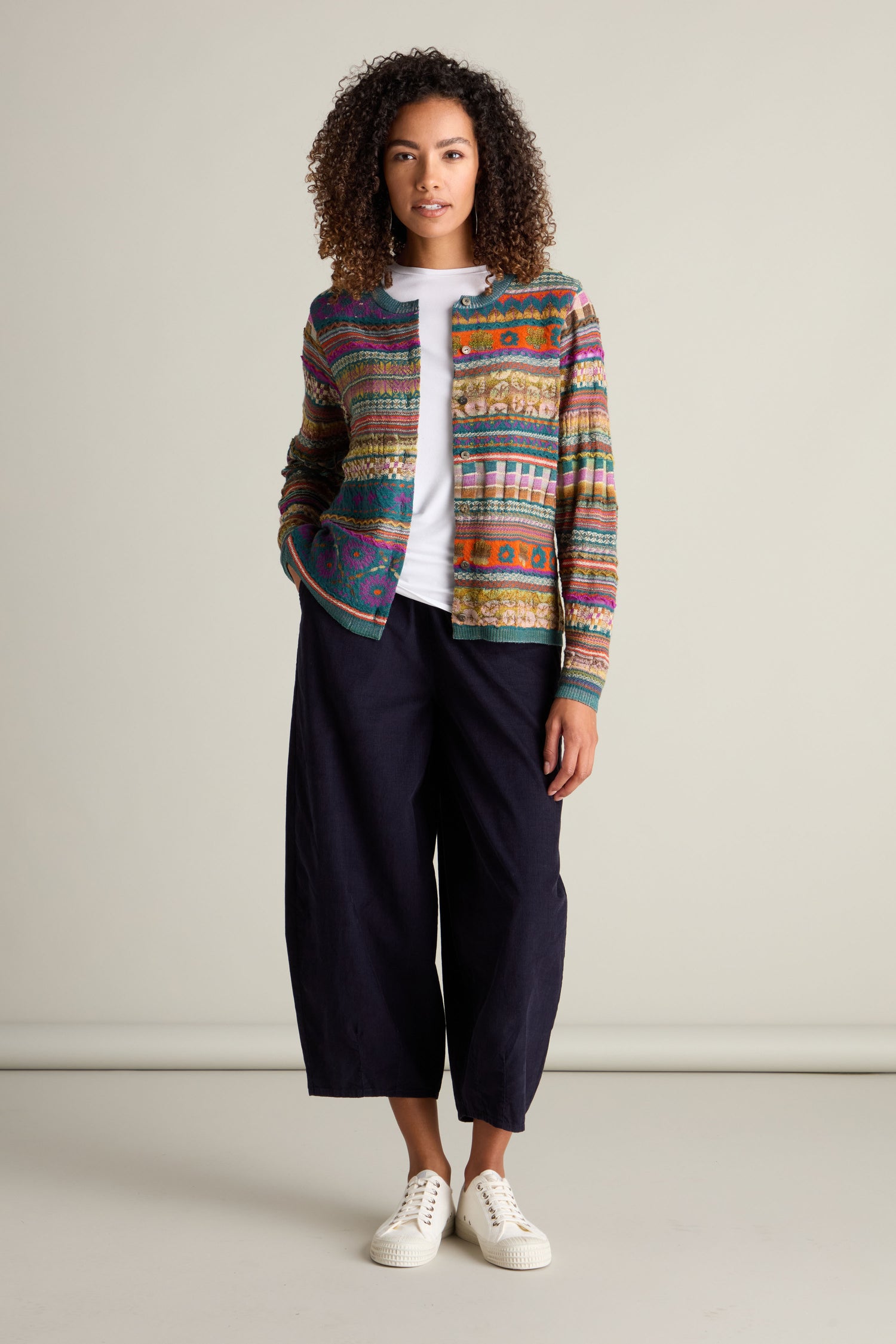 A person stands against a plain background, wearing a colorful Round Neck Button Cardigan with unique patterns over a white shirt, paired with dark loose-fitting pants and white sneakers. Their hands are in the cardigan pockets.