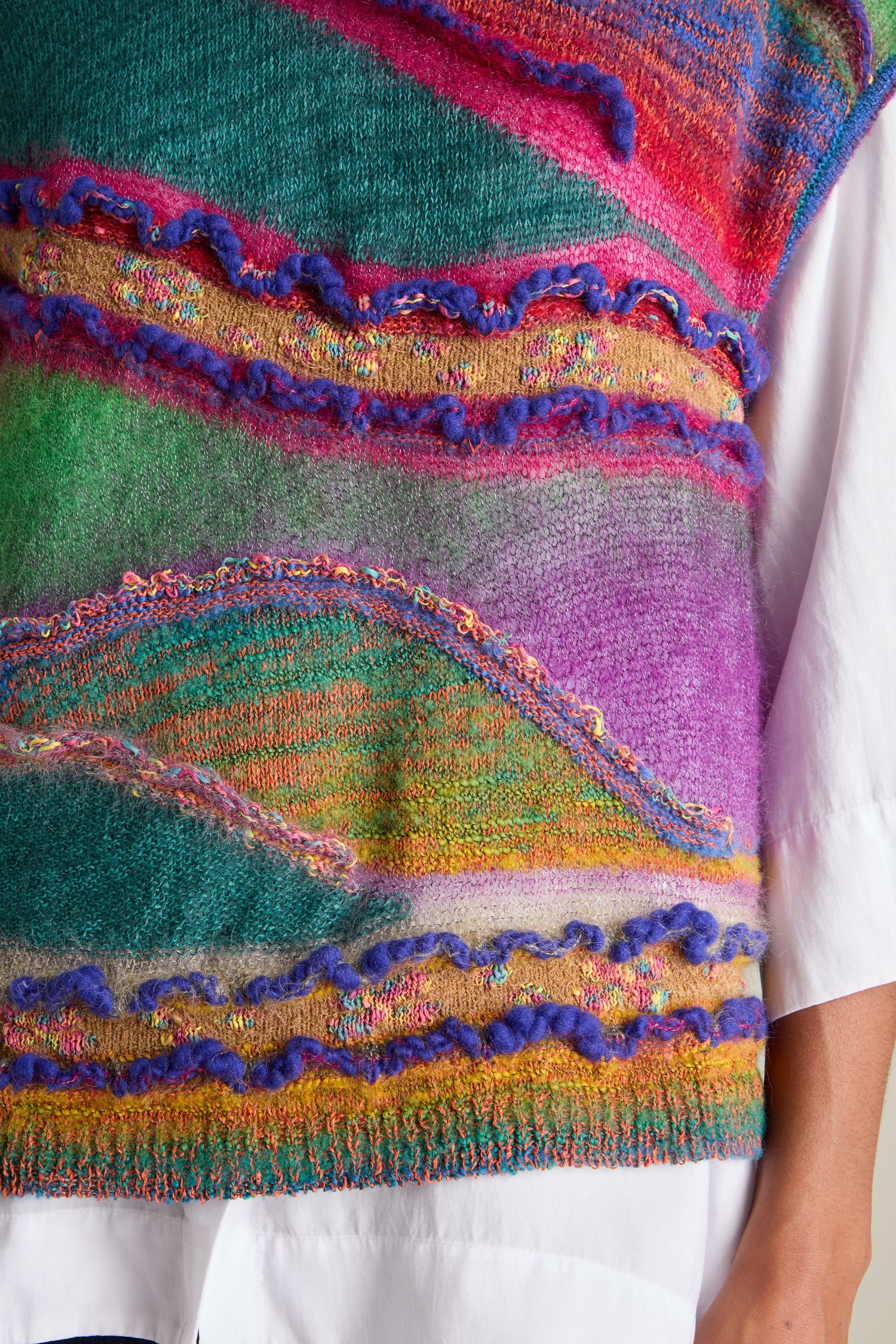 Close-up of the Stitch Detail Tabard, a multicolored knitted sweater featuring wavy patterns over a white button-down shirt. The design showcases vibrant hues of purple, green, pink, and yellow yarns, highlighting intricate stitch details for a touch of modern knitwear elegance.