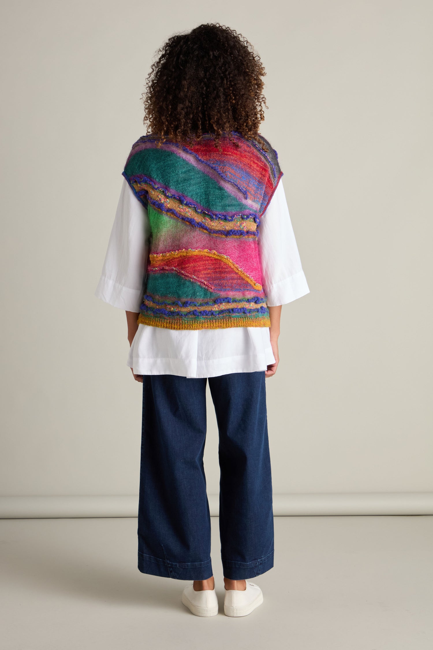 A person with curly hair seen from the back, wearing an eye-catching Stitch Detail Tabard in vibrant hues over a white shirt and blue wide-leg trousers, standing in front of a plain background.