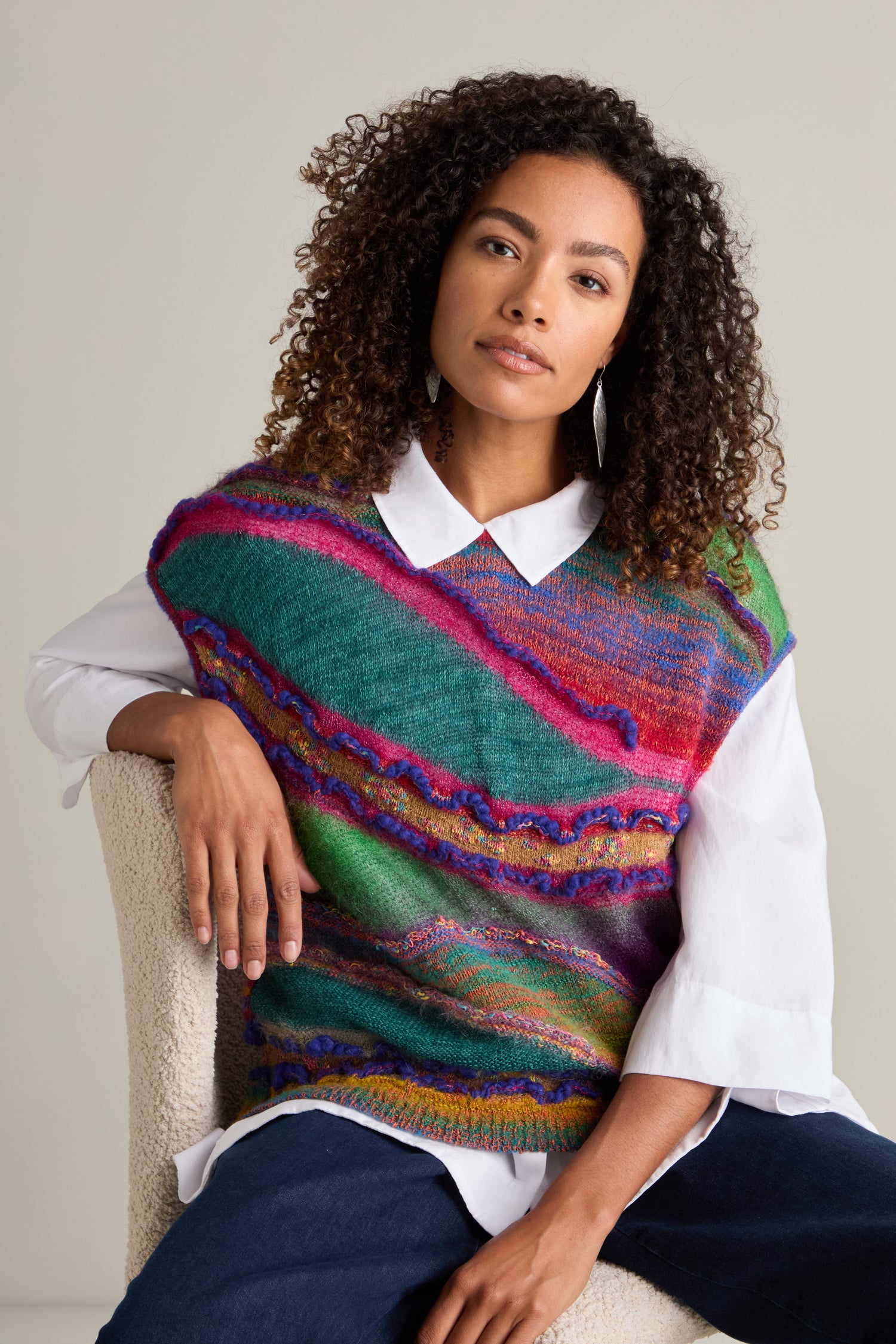 A person with curly hair sits on an off-white chair, wearing a white shirt and a vibrant Stitch Detail Tabard featuring colorful hues.