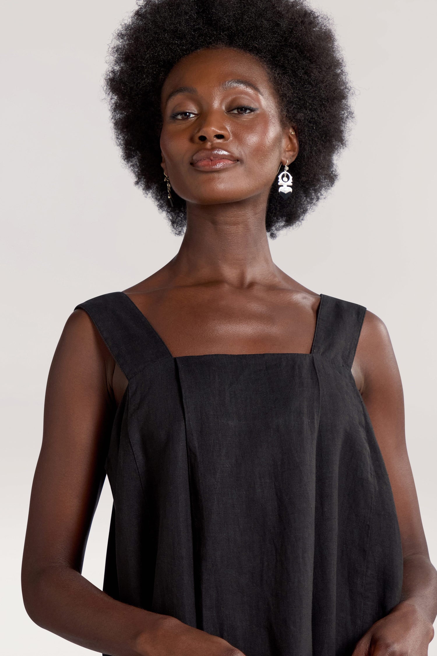 A person with an afro hairstyle is wearing a black sleeveless Square Neck Bubble Linen Dress and earrings, looking confidently at the camera against a neutral background. Perfect for your summer rotation, this Square Neck Bubble Linen Dress adds a touch of elegance to any occasion.