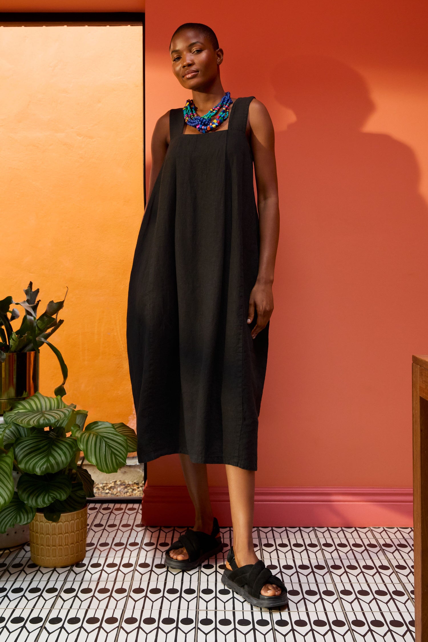 A person stands on a patterned tile floor against an orange wall, wearing a loose black Square Neck Bubble Linen Dress, blue necklace, and black sandals, with one foot forward. Potted plants are nearby.