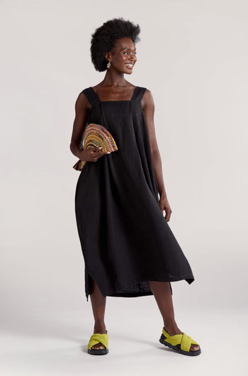 A woman in a black Square Neck Bubble Linen Dress holds a colorful woven clutch and smiles. She wears green sandals and has curly hair. The background is plain and light-colored, making it perfect for her summer rotation.