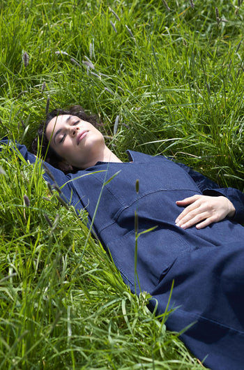 A person wearing a Stretch Denim Pocket Dress is lying in the grass with eyes closed and one hand on their abdomen.