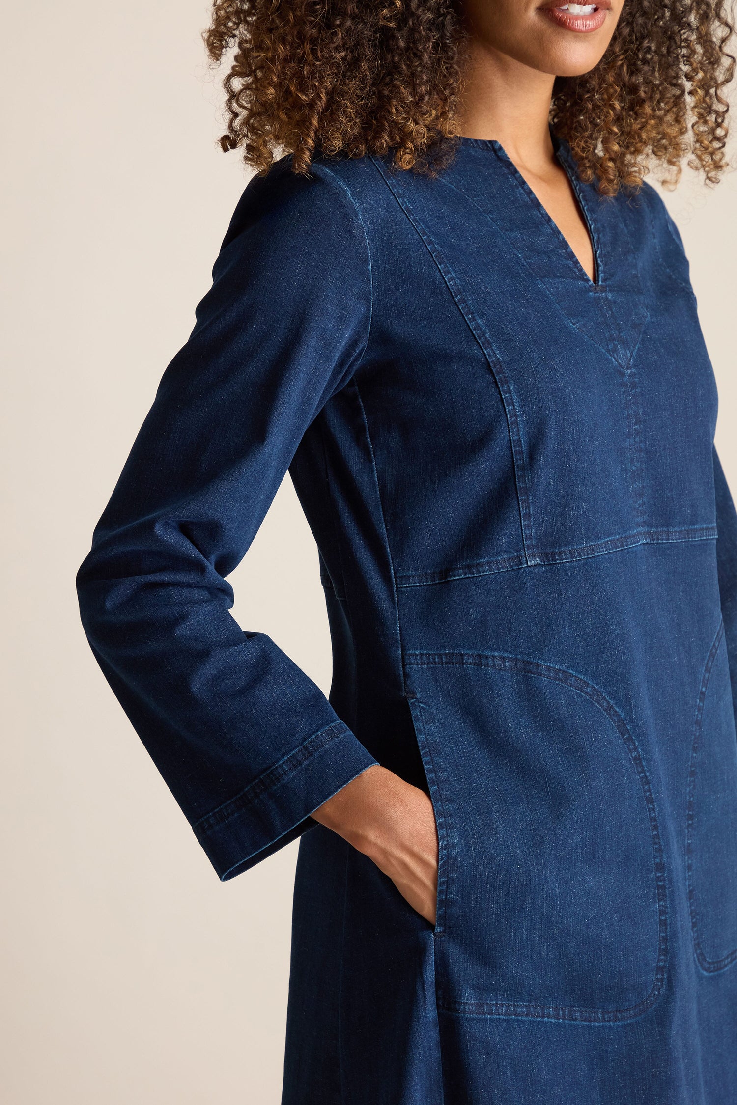 A person with curly hair wears a long-sleeve, dark blue Stretch Denim Pocket Dress featuring an A-line silhouette and a calf-length skirt, standing against a neutral background.