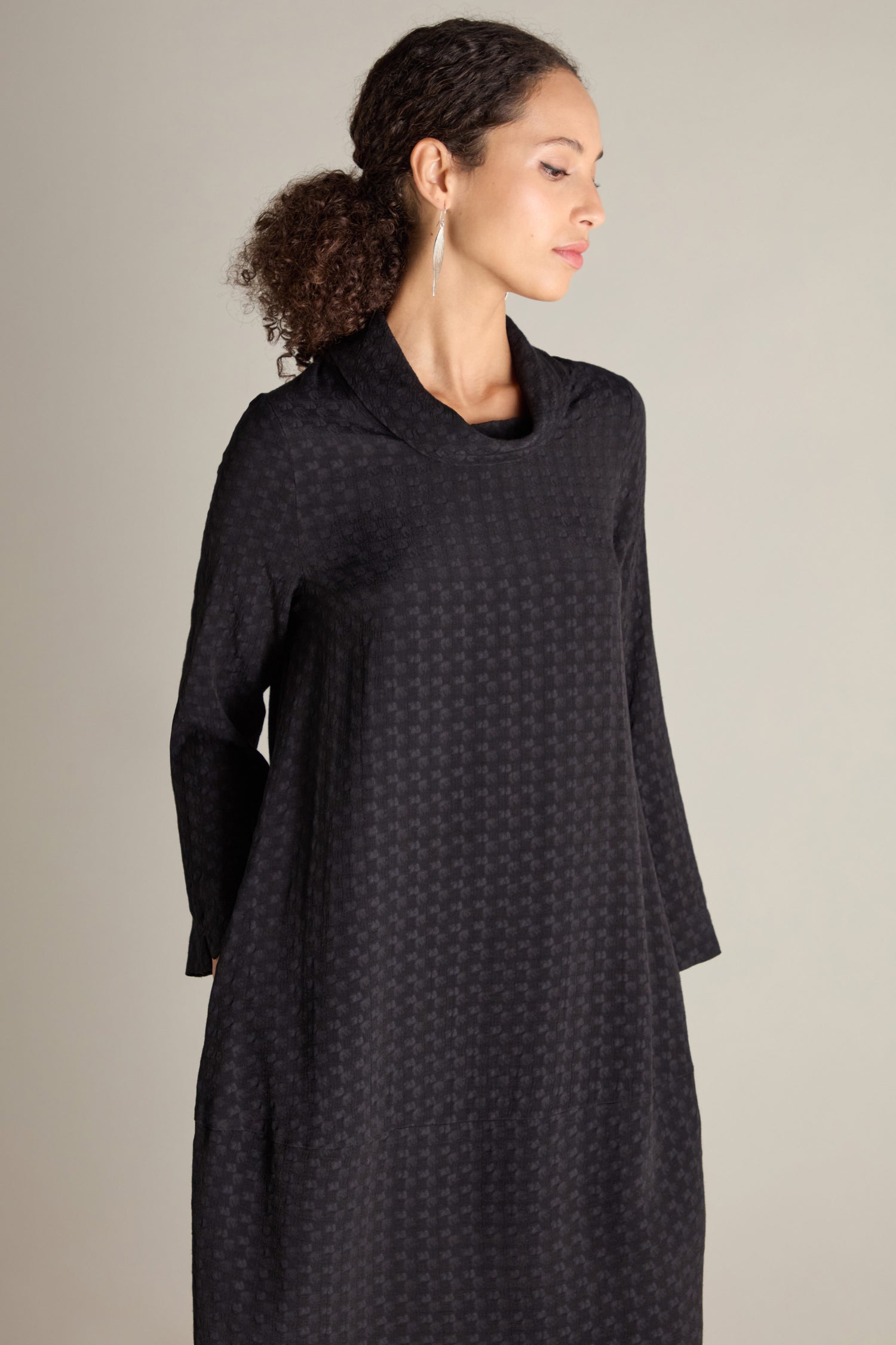 A woman with curly hair pulled back is wearing a long-sleeved, black Small Square Jacquard Bubble Dress that features a voluminous and flattering drape. She is standing in front of a plain, light-colored background.