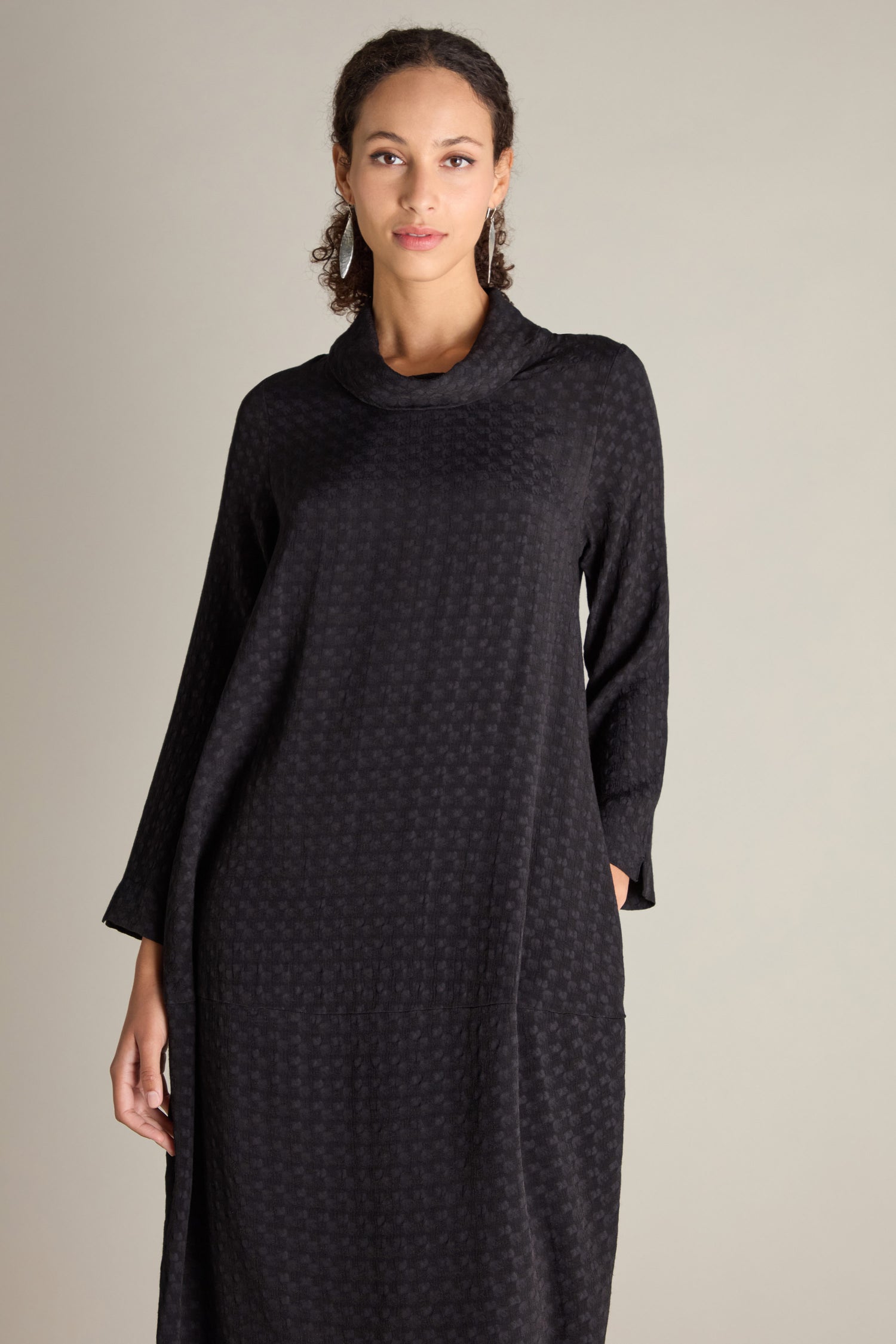 A person with curly hair is standing facing forward, wearing a long-sleeved Small Square Jacquard Bubble Dress in black with a voluminous flattering drape against a plain background.